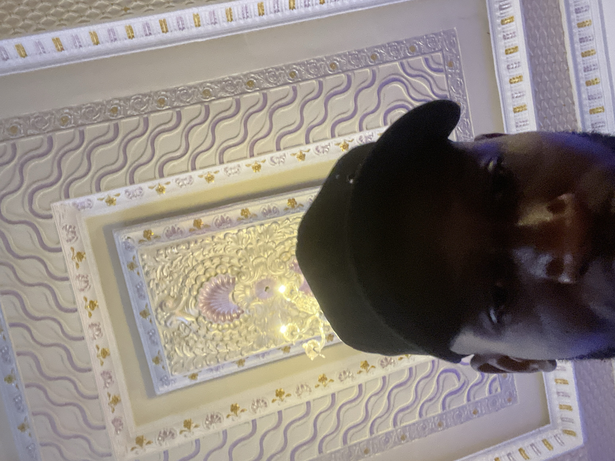 The media file shows a close-up, upside-down shot of a person's face and head, wearing a black baseball cap. The subject is looking upwards at an ornate, white, and gold ceiling. The ceiling features intricate decorative molding with wave-like patterns in purple and white, interspersed with gold floral motifs. In the center of the visible ceiling area is a recessed panel with more detailed ornamentation, including what appears to be a seashell design and a chandelier with warm, glowing lights. The lighting suggests it could be daytime or evening, with the artificial light from the chandelier also illuminating the scene. The perspective of the camera suggests the person is lying down or looking up from a low angle, possibly capturing a selfie or being photographed from below. The overall impression is one of luxury and detail in the indoor architectural setting, contrasted with the casual presence of the person in the foreground. There is no visible text in the image. The location context is Jos, Nigeria.