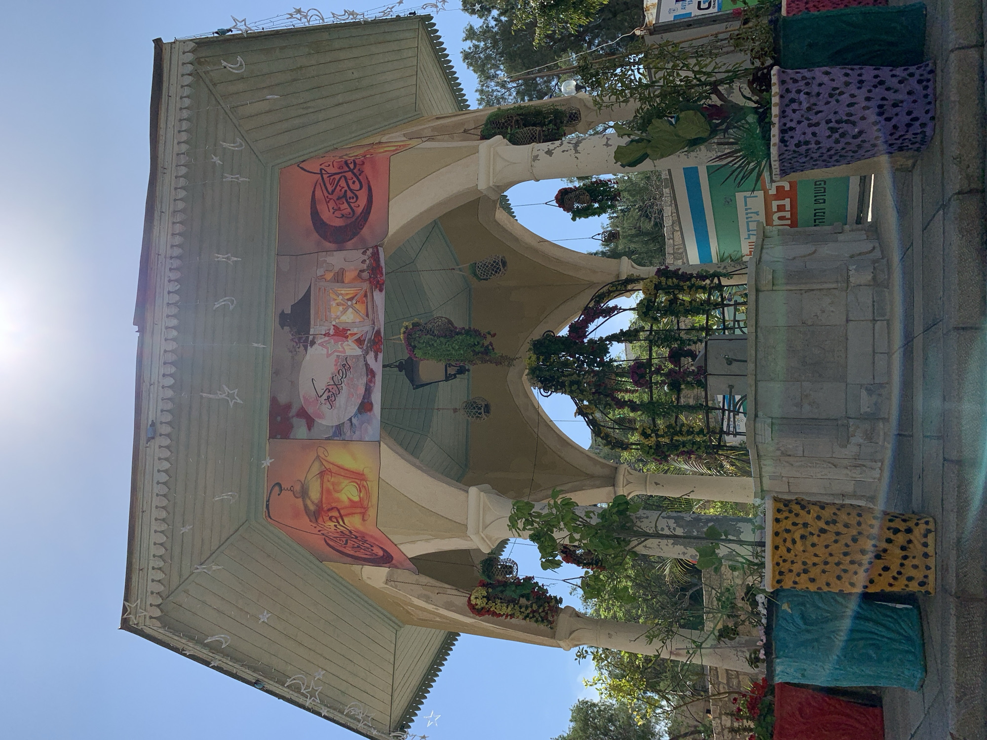 The image depicts a structure with an ornate roof and columns, likely a small pavilion or gazebo. The structure is adorned with images that appear to be themed around a holiday or celebration, possibly Ramadan or a similar cultural event, based on the crescent moon and Arabic calligraphy-style designs. Hanging floral arrangements decorate the columns and arches.
The location is suggested to be in Tel Aviv, Israel. The style of architecture and the cultural elements in the images suggest that the structure could be located in a historic or cultural district of the city, possibly Jaffa or Neve Tzedek.
The scene is captured during the day, with clear blue skies visible above the structure. There are planter boxes with vibrant colors and patterns positioned along the edges of the structure, adding to the festive atmosphere. The image appears to be taken from a low angle, emphasizing the height and architectural details of the structure.