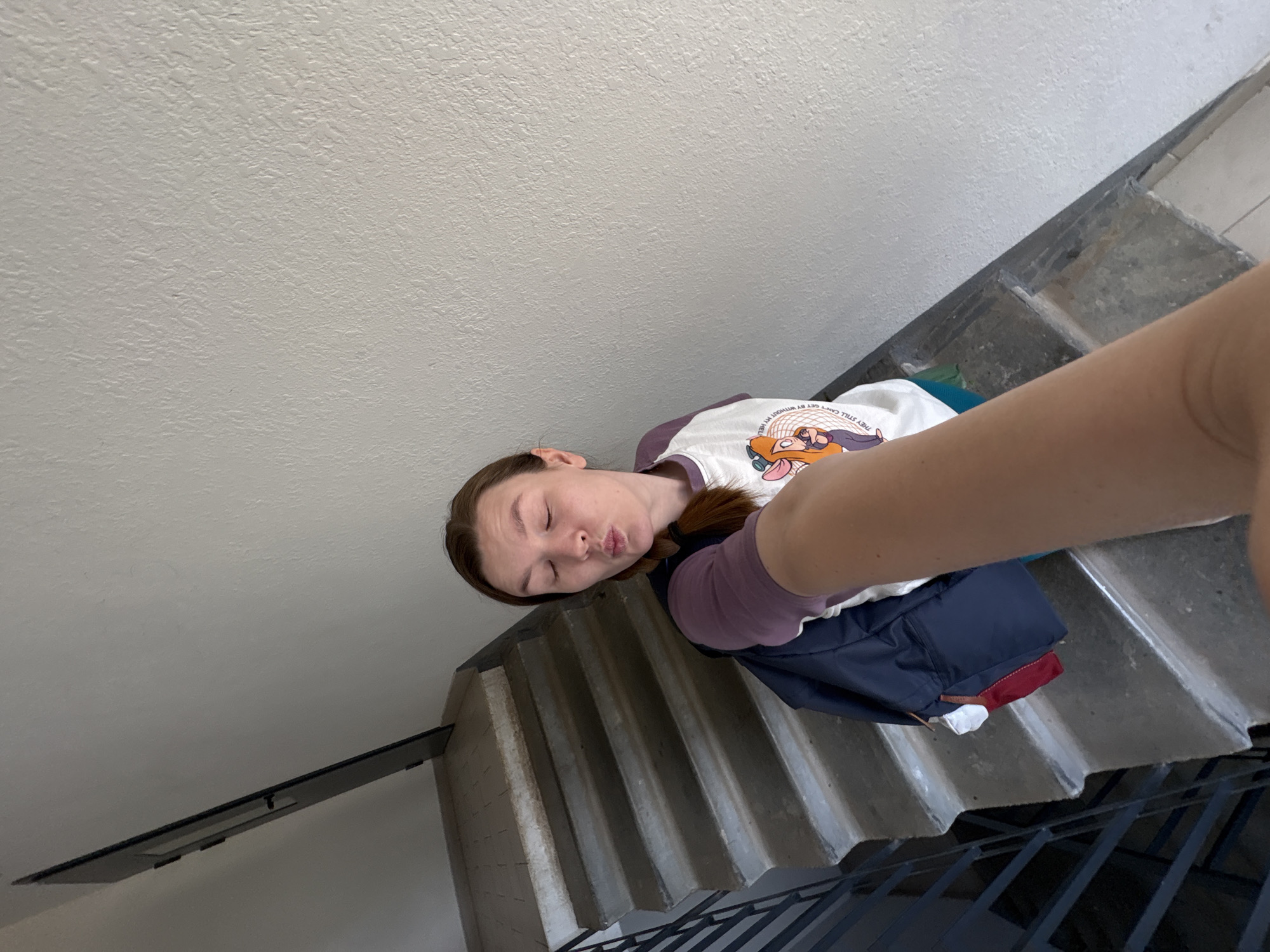 A high-angle selfie photograph depicts a woman posing in an interior stairwell. The woman, positioned in the mid-right of the frame, has light skin, brown hair styled in a braid over her left shoulder, and her eyes closed with lips pursed in an exaggerated expression. She is wearing a long-sleeved shirt with purple sleeves and a white torso panel featuring a graphic of animated cat characters and partial text. A dark blue backpack, from which a red fabric item and a white item are visible, is worn over her left shoulder. Her right arm is extended, holding the camera to capture the image, with her forearm partially obscuring the lower right foreground.
The background reveals a concrete staircase ascending from the lower left to the upper right. The steps exhibit wear, dirt, and scuff marks, with eroded light-colored nosings. A dark metal railing with vertical bars parallels the left side of the staircase. The wall to the right of the woman and the stairs is light-colored, off-white or light grey, and displays a prominent rough texture, indicative of stucco or textured paint. The wall on the left side of the staircase appears darker due to shadow. The environment is utilitarian, consistent with a common area stairwell in a building. The image contains no discernible visual elements that could confirm its location in Izhevsk, Russia.