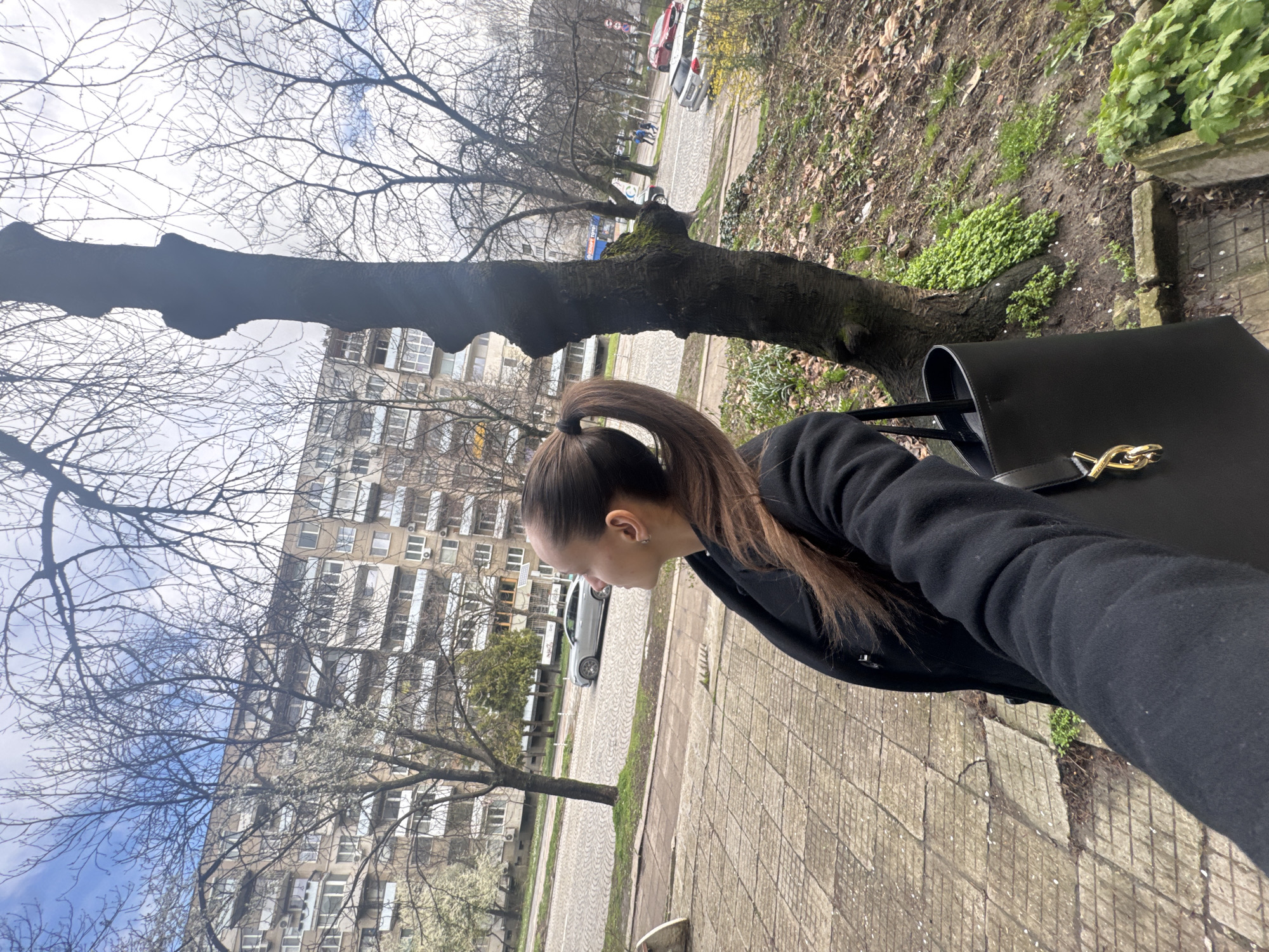 The image shows a woman standing on a sidewalk in Sofia, Bulgaria. She has brown hair pulled back in a ponytail and is wearing a black jacket. She is carrying a large black handbag. Behind her is a street with cars parked along the side. In the background, there is a large apartment building with multiple windows. The trees are bare with just a few blossoms starting to show. The sidewalk is paved with gray bricks.