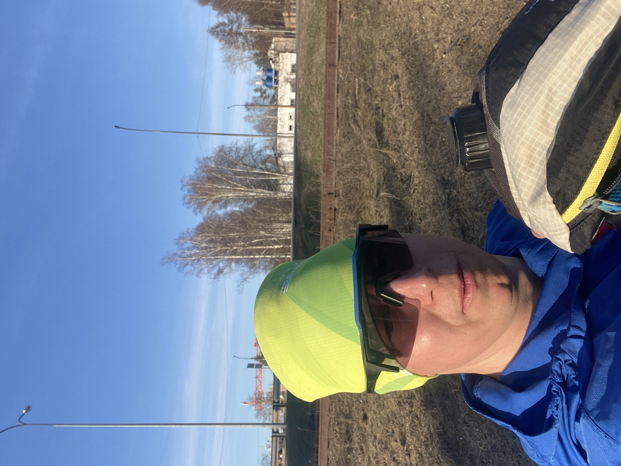 A person wearing a blue hooded jacket, a yellow and green cap, and dark sunglasses is taking a selfie outdoors. The background shows a clear blue sky, bare trees, streetlights, and a building in the distance. A crane is visible amongst the trees. The person is also wearing a backpack. The photo appears to be taken near a railway track. It is difficult to confirm the location as Chaikovsky, Russia, without additional contextual clues.