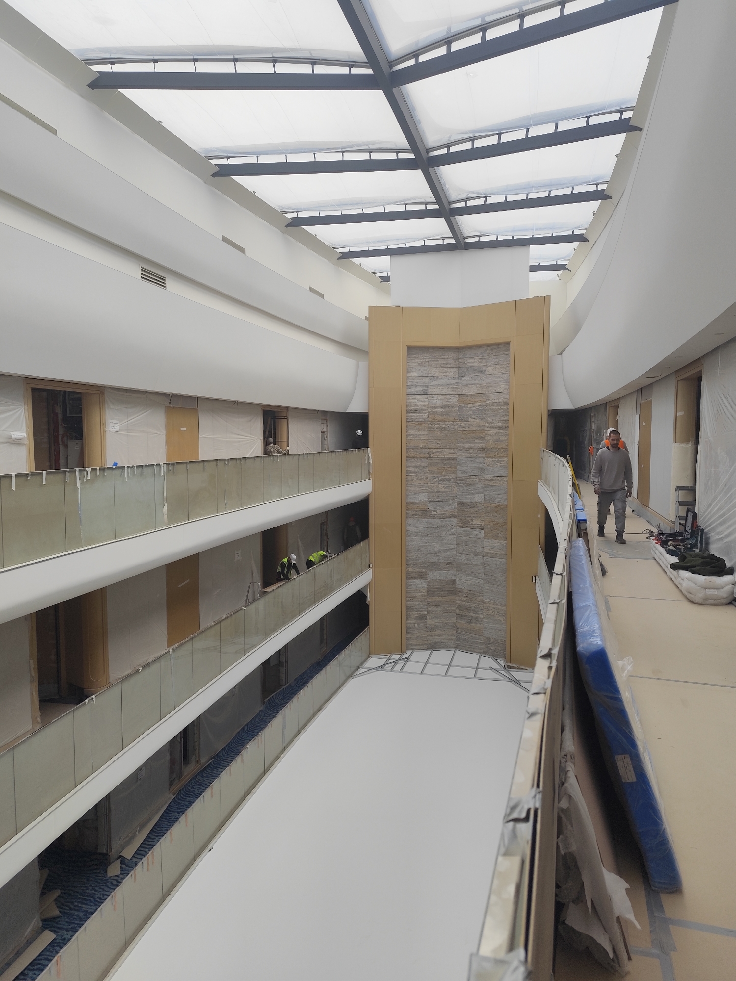 The image depicts the interior of a multi-story atrium-style building, possibly a hotel or large residential complex, in an unknown city and unknown location. The architectural style is modern, with a high ceiling featuring a grid-like skylight structure. The building appears to be under construction or renovation, as evidenced by the presence of protective coverings on walls, furniture, and fixtures.
There are at least three levels visible, with railings along the open spaces overlooking the atrium. A decorative feature, possibly a stone veneer wall, is located in the center of the atrium.
At least three individuals are visible. One man, wearing what appears to be a construction worker's uniform, is walking along a hallway on one of the upper levels. There are at least two additional workers visible from a lower level. Tools and materials are scattered along the hallway, indicating active construction or renovation work. The environment is bright and airy due to the natural light from the skylight.