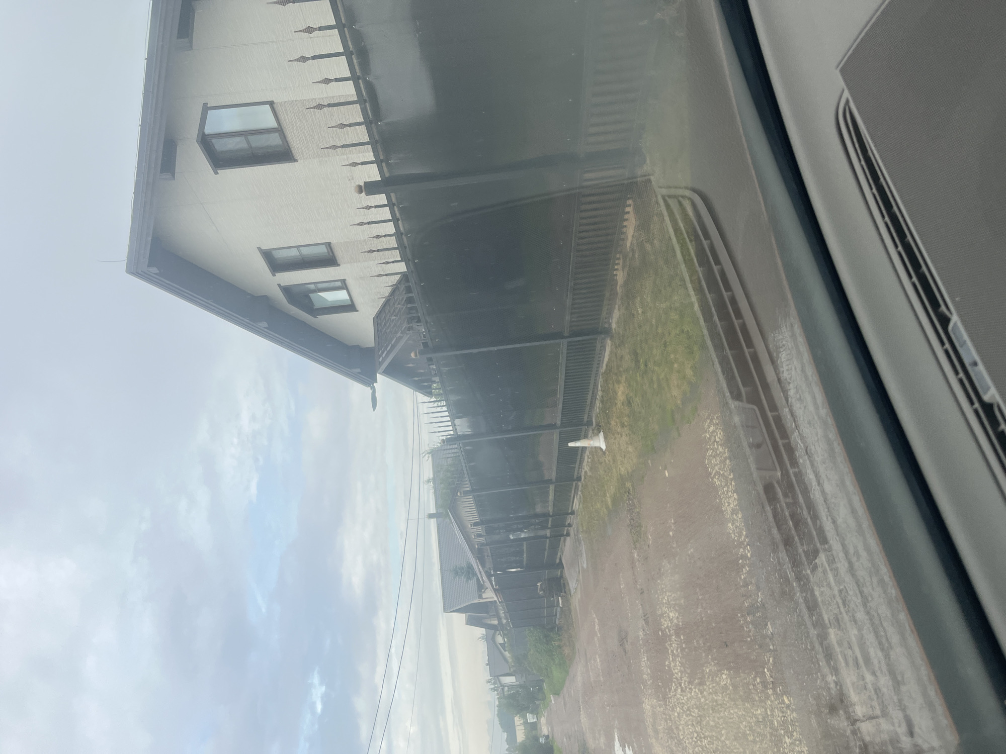 The image shows a street in Vladimirovka, Russia, with several houses and a tall, dark fence. The road appears to be unpaved and wet, with some puddles visible. The sky is overcast, suggesting a cloudy or rainy day.
In the foreground, the dashboard of a vehicle is visible, indicating the photo was taken from inside a car. The fence runs along the right side of the street, separating the road from the properties behind it. The fence is made of dark metal with decorative spikes along the top.
Behind the fence, a light-colored, multi-story house with a gabled roof is visible. It has several windows, some of which are bay windows. Further down the street, more houses can be seen, as well as trees and vegetation. A white traffic cone is placed on the side of the road near the fence. Overhead power lines are also visible against the sky. The overall impression is of a residential area on a day with subdued lighting conditions.