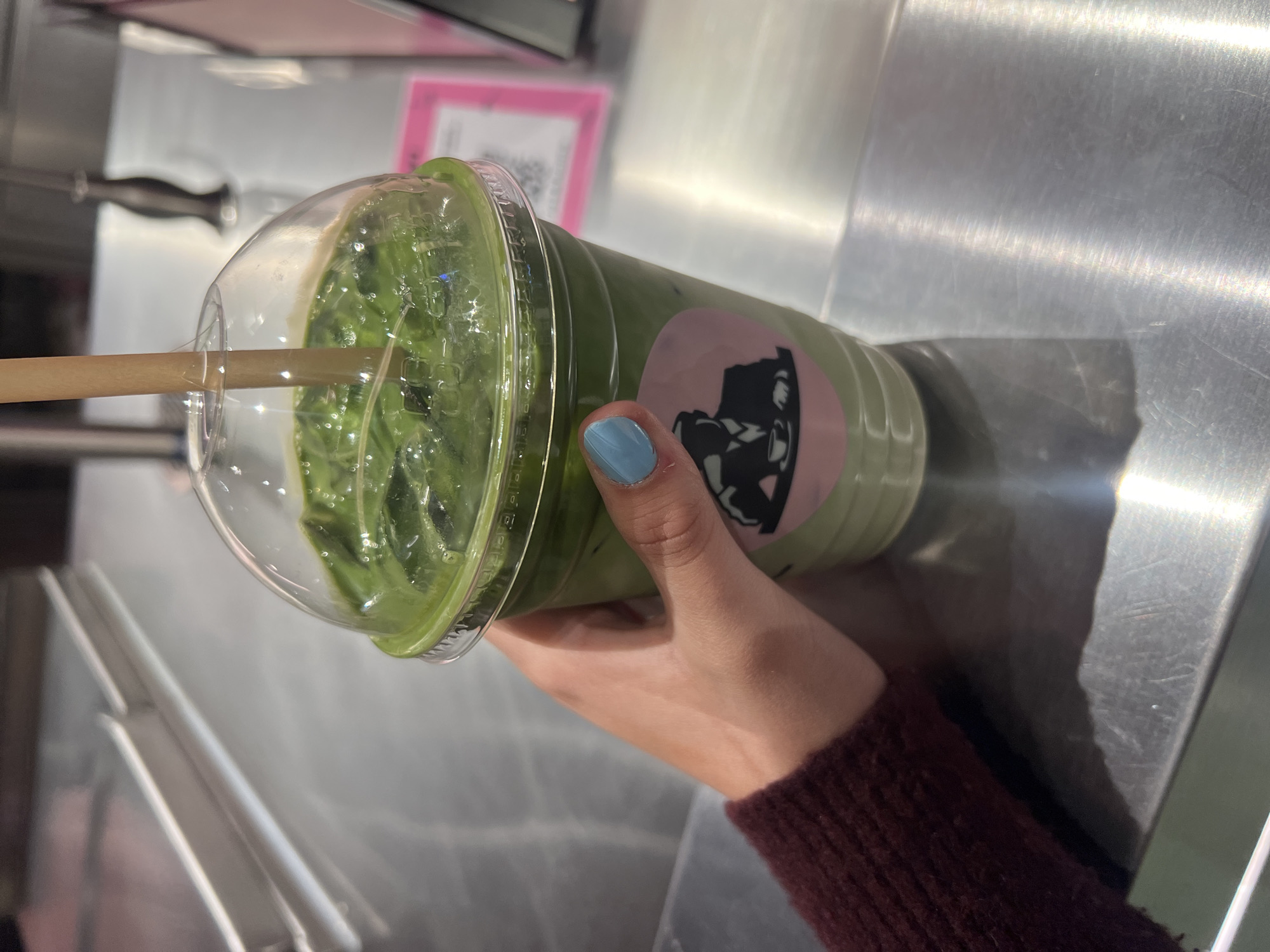 A person is holding a clear plastic cup filled with a green beverage and ice, topped with a dome lid and a brown straw. The drink is held in front of a metallic surface with visible reflections. The person's hand has light blue nail polish. The person is wearing a maroon sweater. A pink sticker or sign is visible in the background behind the cup. The location appears to be indoors, possibly a cafe or restaurant, as suggested by the metallic surfaces and background elements. The location is Unknown, Unknown.