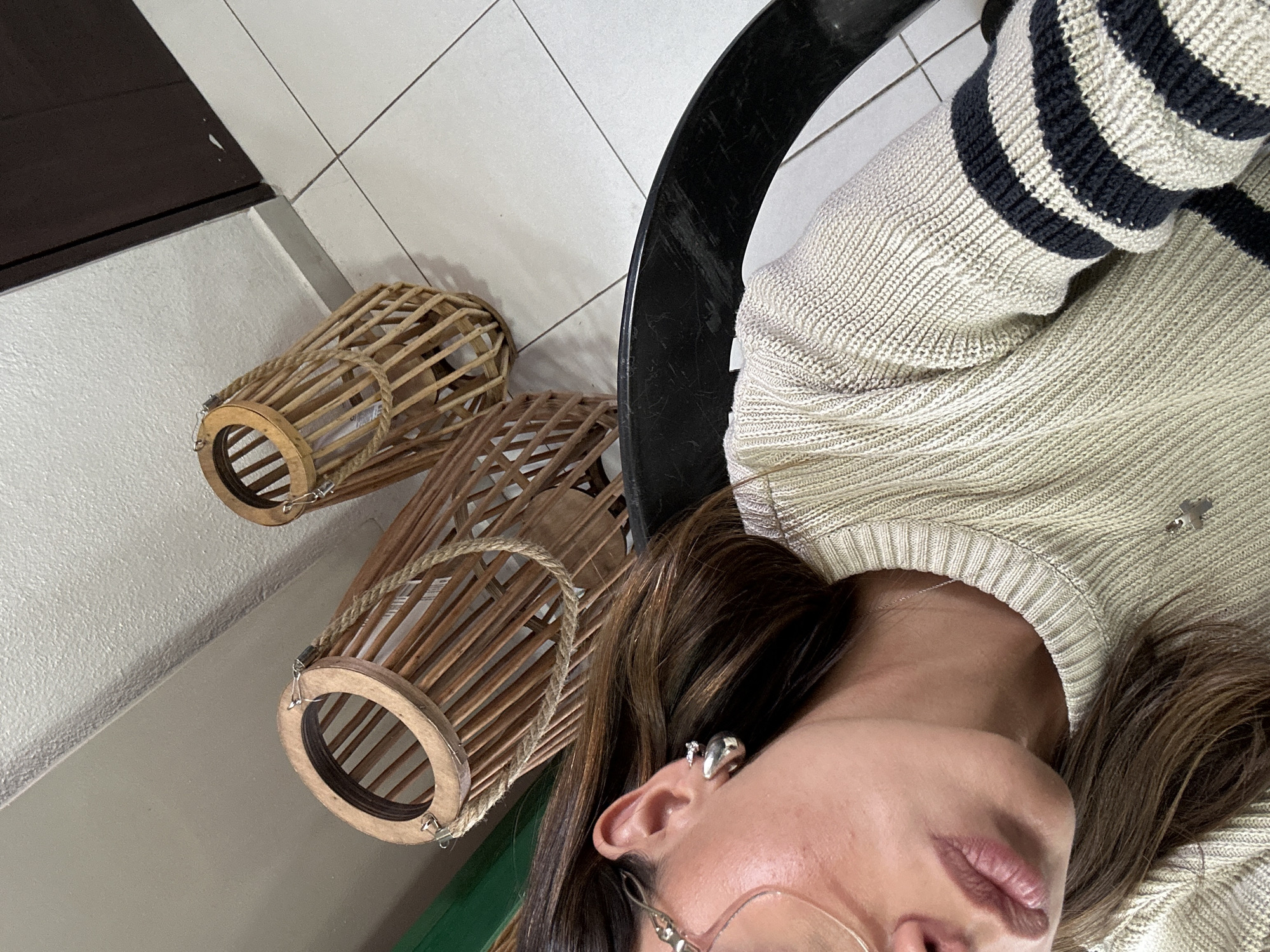 A close-up, tilted photograph shows a person wearing a beige and navy striped sweater, resting against a black chair back. Adjacent to the chair are two wooden, cylindrical lanterns of different sizes, placed on a surface that appears to be a wall or floor painted in shades of green, beige, and white. The person in the photograph has brown hair, is wearing earrings, and has eyeglasses. The location is Unknown, Unknown.