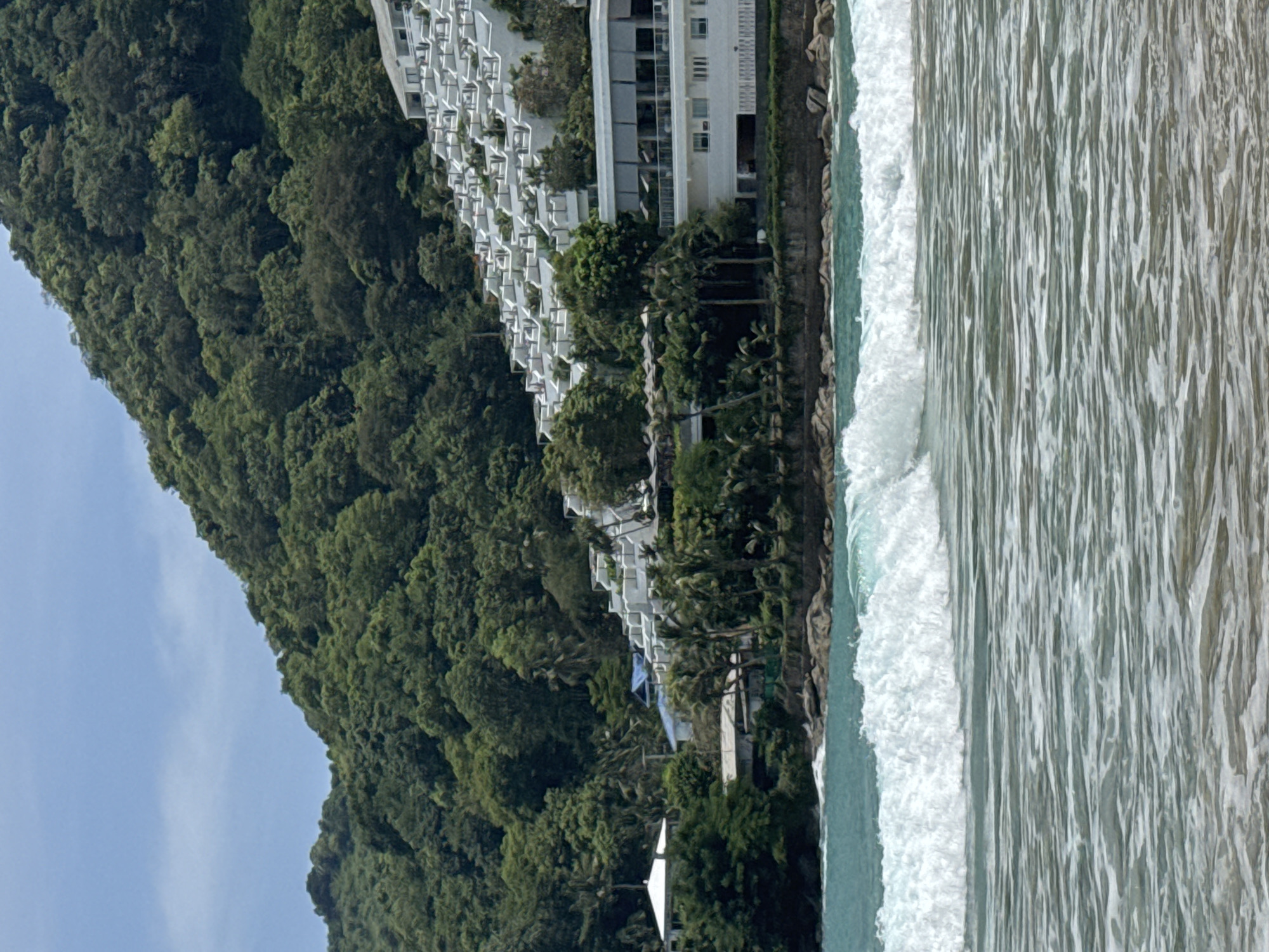 The image captures a coastal scene in Rawai, Thailand, characterized by a lush green hillside densely covered with trees. Cascading down the hillside is a multi-tiered white building complex, appearing to be a resort or hotel, with numerous balconies and white railings. The foreground is dominated by the ocean, with a large, foamy wave cresting and breaking. The water transitions from a turquoise hue near the shore to a lighter, rippled surface with white foam further out. Several rocks are visible along the shoreline. A few palm trees are interspersed amongst the vegetation on the hillside, and a couple of smaller structures are visible at the base of the hill, closer to the water. The sky above is a pale blue with a few wispy clouds.