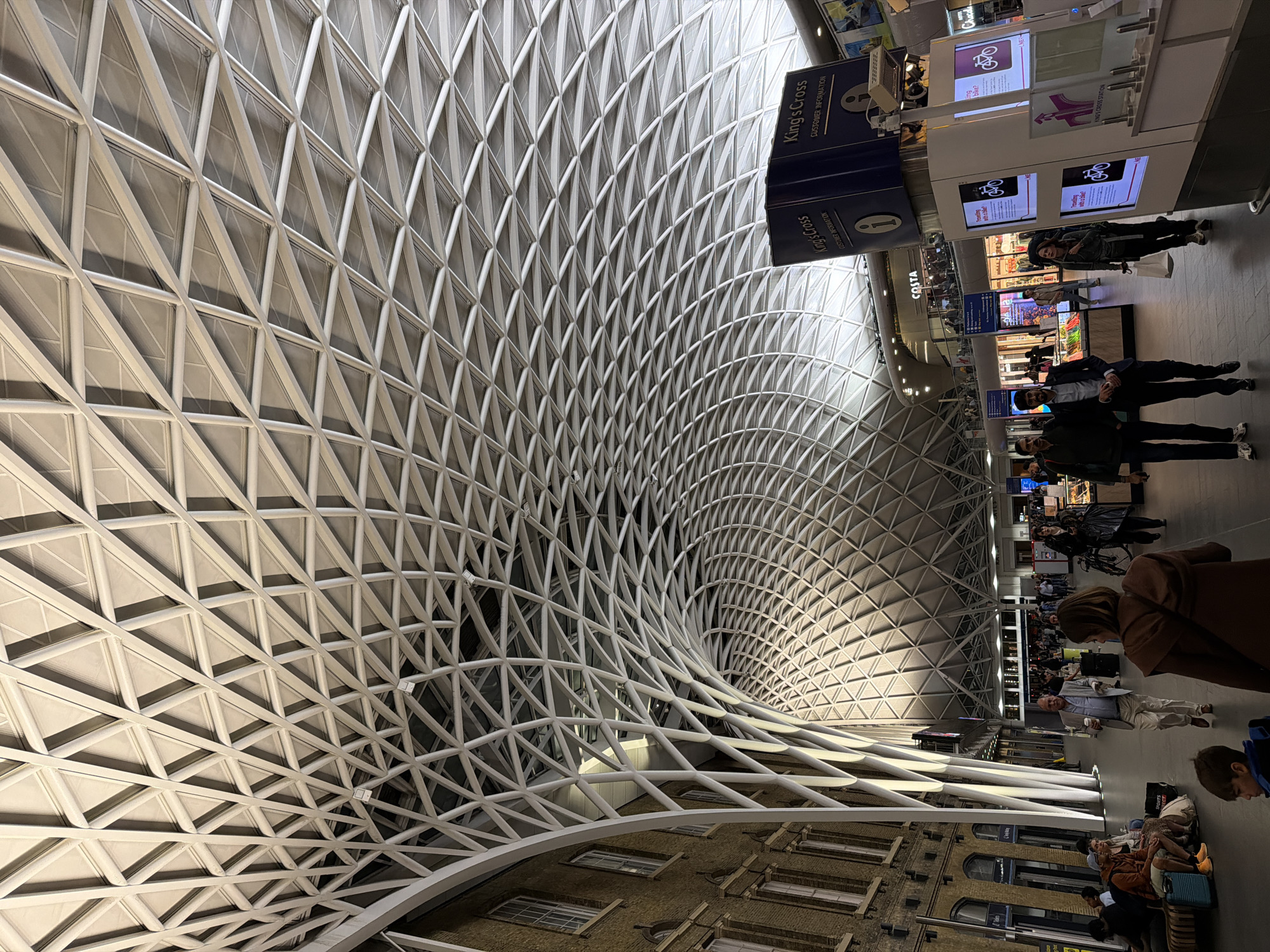 The image depicts the interior of King's Cross Station in London, United Kingdom. The focus is on the station's distinctive roof structure, a complex web of interconnected triangular panels made of a light-colored material. The roof curves dramatically overhead, creating a sense of vastness.
The station's concourse is populated with people walking, standing, and sitting. There are signs visible, one indicating "King's Cross Customer Information" and another for "Costa." There are also digital displays showing various advertisements and public service announcements. The lower part of the image shows the exterior of a building.