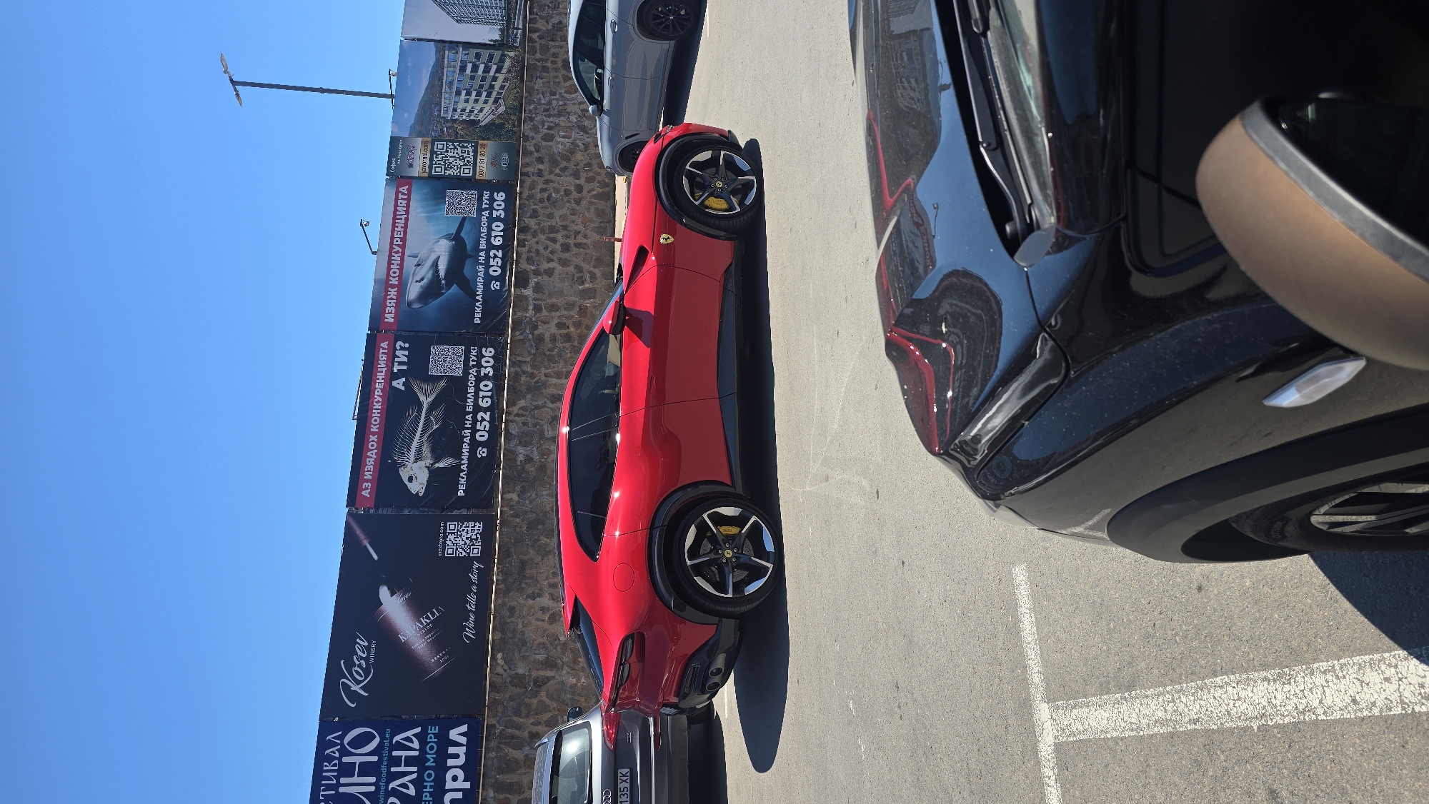 The image captures a bright, sunny day in Varna, Bulgaria, likely in a parking lot. The foreground shows the hood and side mirror of a black car, reflecting the red car parked in front. Dominating the mid-ground is a bright red Ferrari Purosangue, parked on asphalt with visible white parking lines. The car is viewed from the side, highlighting its sporty design, unique headlights, and black wheels with yellow brake calipers. Behind the Ferrari, a grey SUV is partially visible. The background features a stone wall topped with a series of large billboards advertising various businesses. One billboard prominently displays a bottle of KOSEV winery with the text "Wine tells a story". Other billboards contain images of a fish skeleton and a shark, along with phone numbers and QR codes. A lamppost stands tall against a clear blue sky, with buildings visible in the far distance.