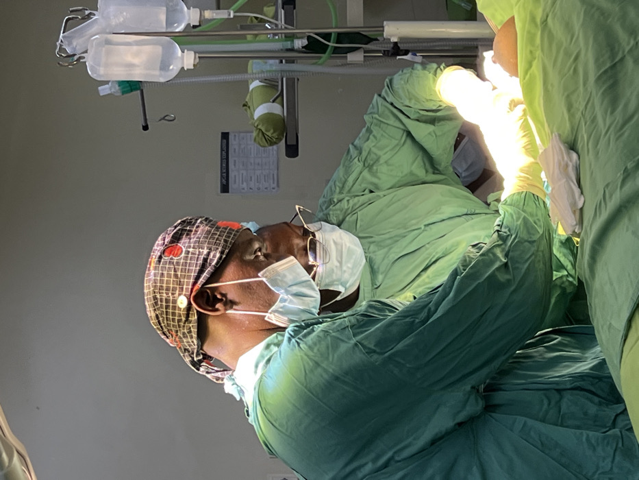 A surgical scene is captured in an operating room within Nairobi, Kenya. Two medical professionals, both wearing green surgical scrubs, are focused on a patient positioned centrally. The individual on the left wears a patterned surgical cap and a surgical mask hanging below their chin, while leaning over the patient. The individual on the right wears a surgical mask covering their mouth and nose, eyeglasses, and has gloved hands illuminated by a bright surgical light, actively engaged with the patient's exposed body part, which is partially covered by green surgical drapes. Unused white surgical gloves rest on the drapes in the foreground. In the background, an IV pole supports two hanging IV bags, and a wall-mounted poster with text partially legible as "APGAR SCORE EXPLAINED" is visible above a shelf holding a rolled green fabric item.