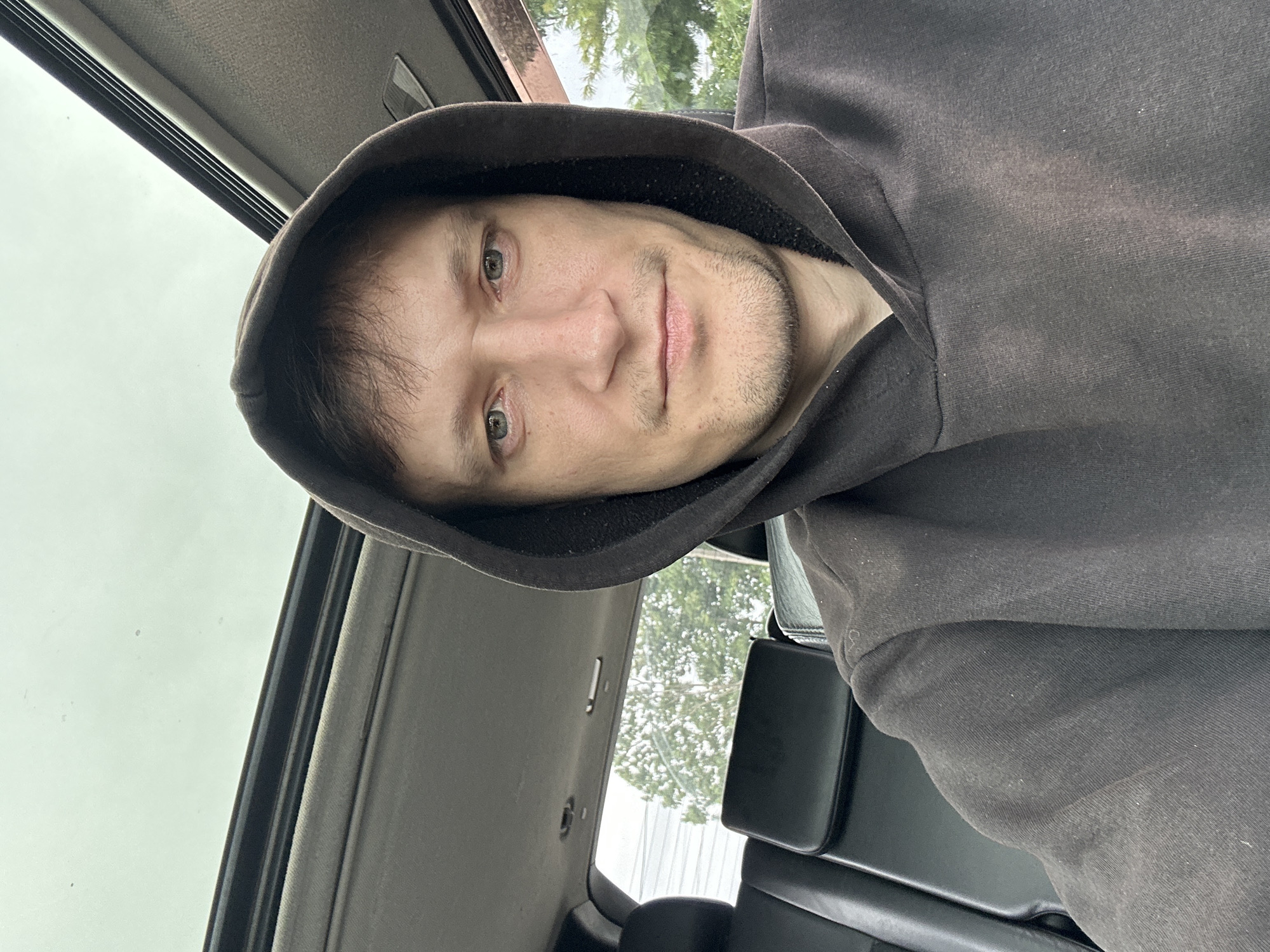 The image is a close-up selfie of a man in a dark hoodie. He is inside a vehicle, with a sunroof open above him. The interior of the car is visible, including black leather seats. Outside the car, trees and power lines can be seen, suggesting an outdoor, likely suburban or rural, setting. The man has fair skin, light-colored eyes, and short brown hair visible under the hood. He has light stubble on his chin and upper lip. The lighting is natural, coming from above and the side. There is no information in the image to suggest the location is the city of Makiv, Ukraine.