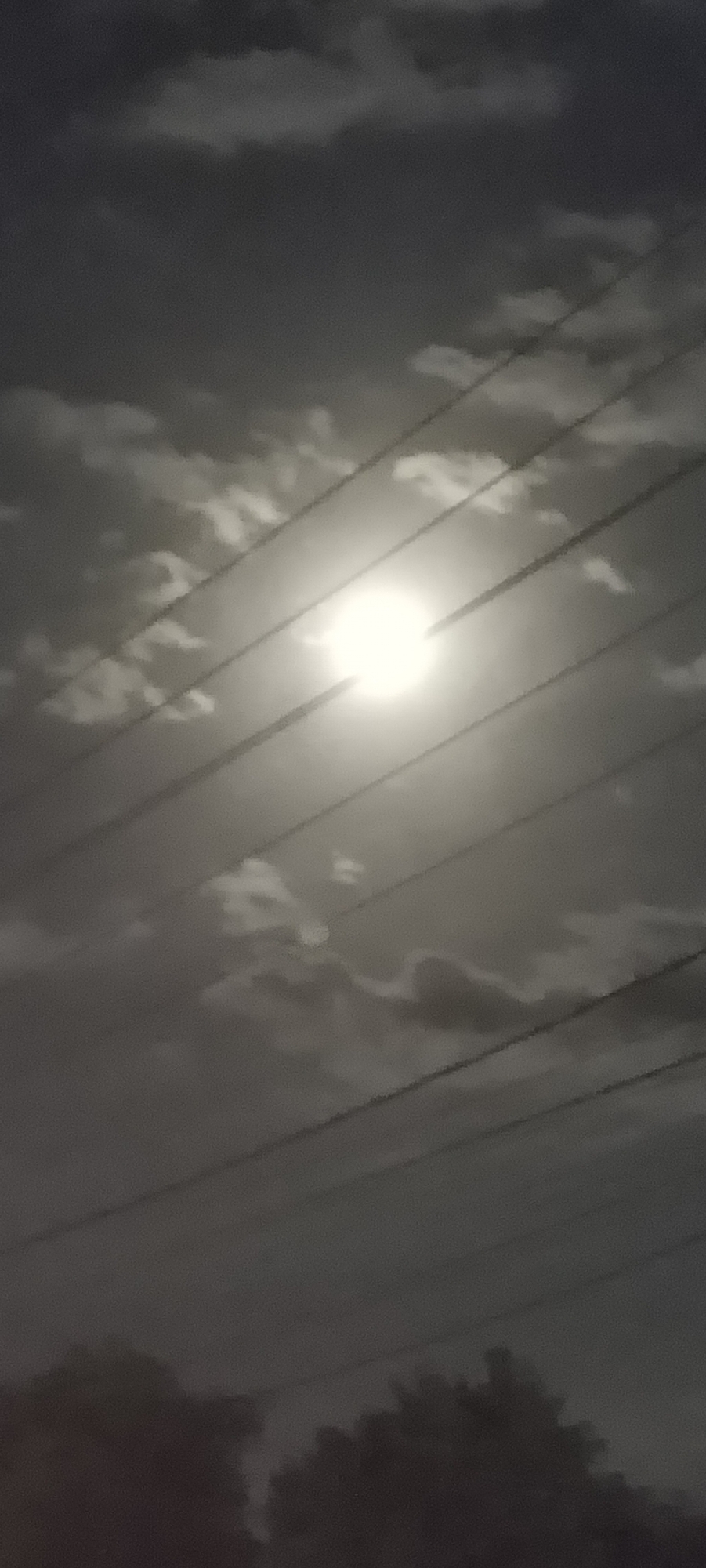 The image shows a night scene, possibly in Meuse, Indonesia, though this cannot be confirmed with certainty based on the image alone. A bright light, likely the moon, dominates the center of the frame, diffused by thin clouds. Power lines run diagonally across the view, partially obscuring the light source. The lower portion of the image suggests trees or foliage in silhouette against the dark sky. The overall impression is one of darkness with a strong, soft light source, and potentially urban or suburban features (power lines).