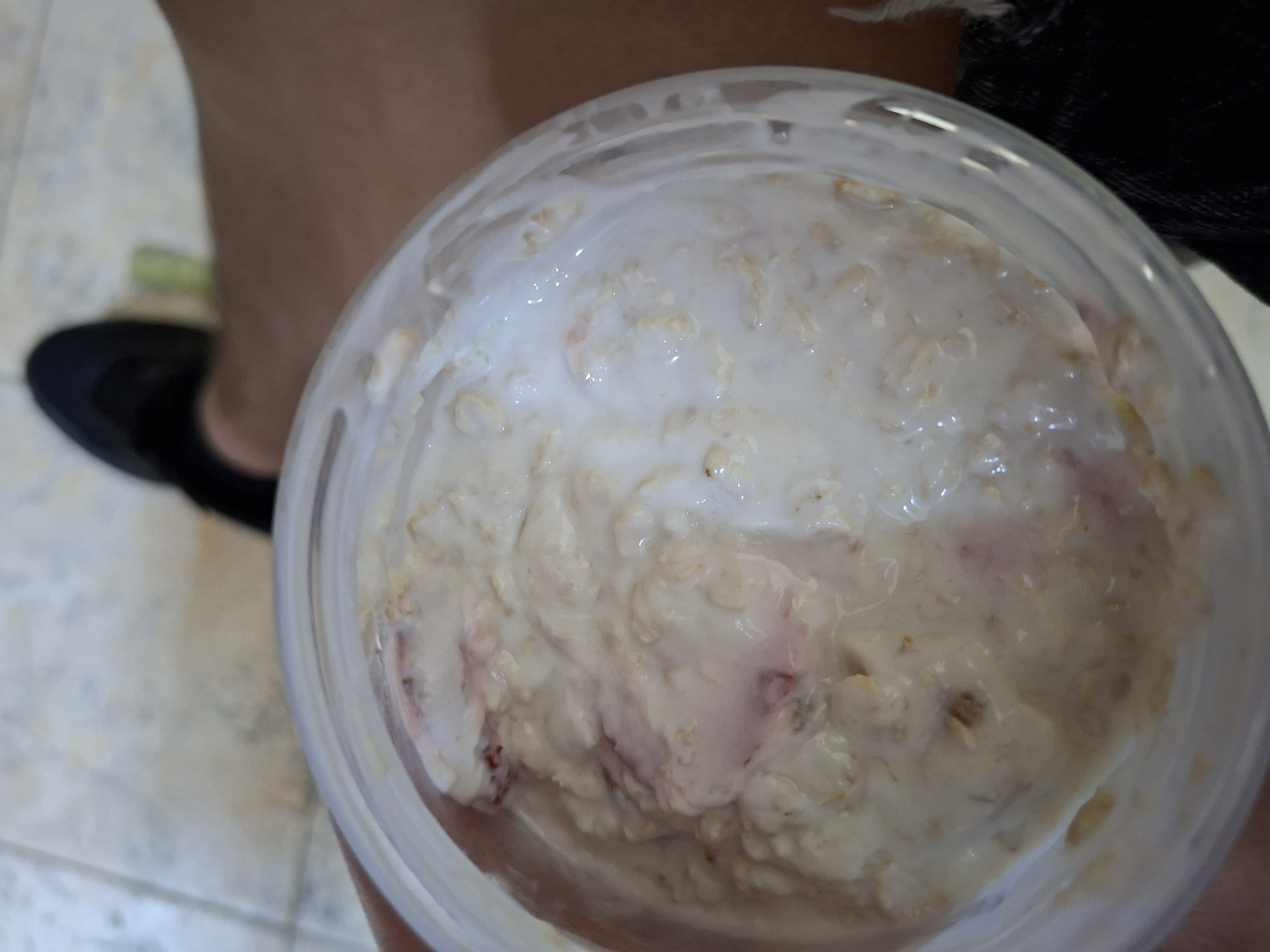 The image captures a top-down view of a clear plastic container filled with a creamy food mixture, likely overnight oats or a similar breakfast/snack, in Ruse, Bulgaria. The contents are predominantly off-white to light beige, with discernible oat flakes and streaks of pink or light red, suggesting the inclusion of berries. A smooth, white, and slightly translucent liquid, possibly milk or yogurt, floats on the surface of the mixture.
In the blurred background, to the left, the lower part of a person's leg and a foot wearing a dark, flat shoe are visible, indicating someone is holding or is near the container. A dark fabric, possibly clothing, can be seen in the upper right background. The floor beneath appears to be a light-colored tiled surface. The scene suggests a casual, indoor setting, likely portraying a moment before the food item is consumed. No text is visible in the image.