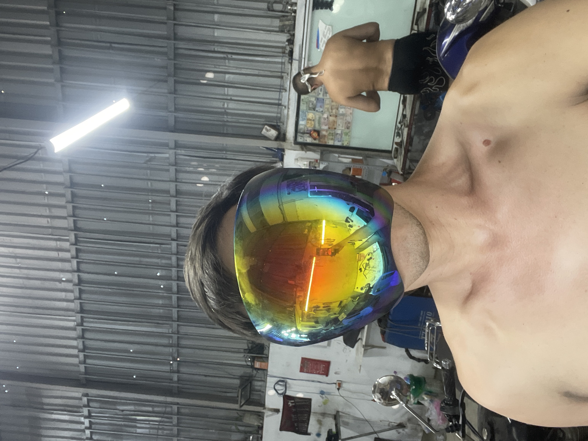 The image shows a close-up of a shirtless man wearing iridescent goggles. The environment appears to be a garage or workshop with exposed metal walls and fluorescent lighting. In the background, another shirtless man with a bandaged head is visible near a display case with money. A motorcycle is nearby. The image was taken in Bac Nha Trang Ward, Vietnam.