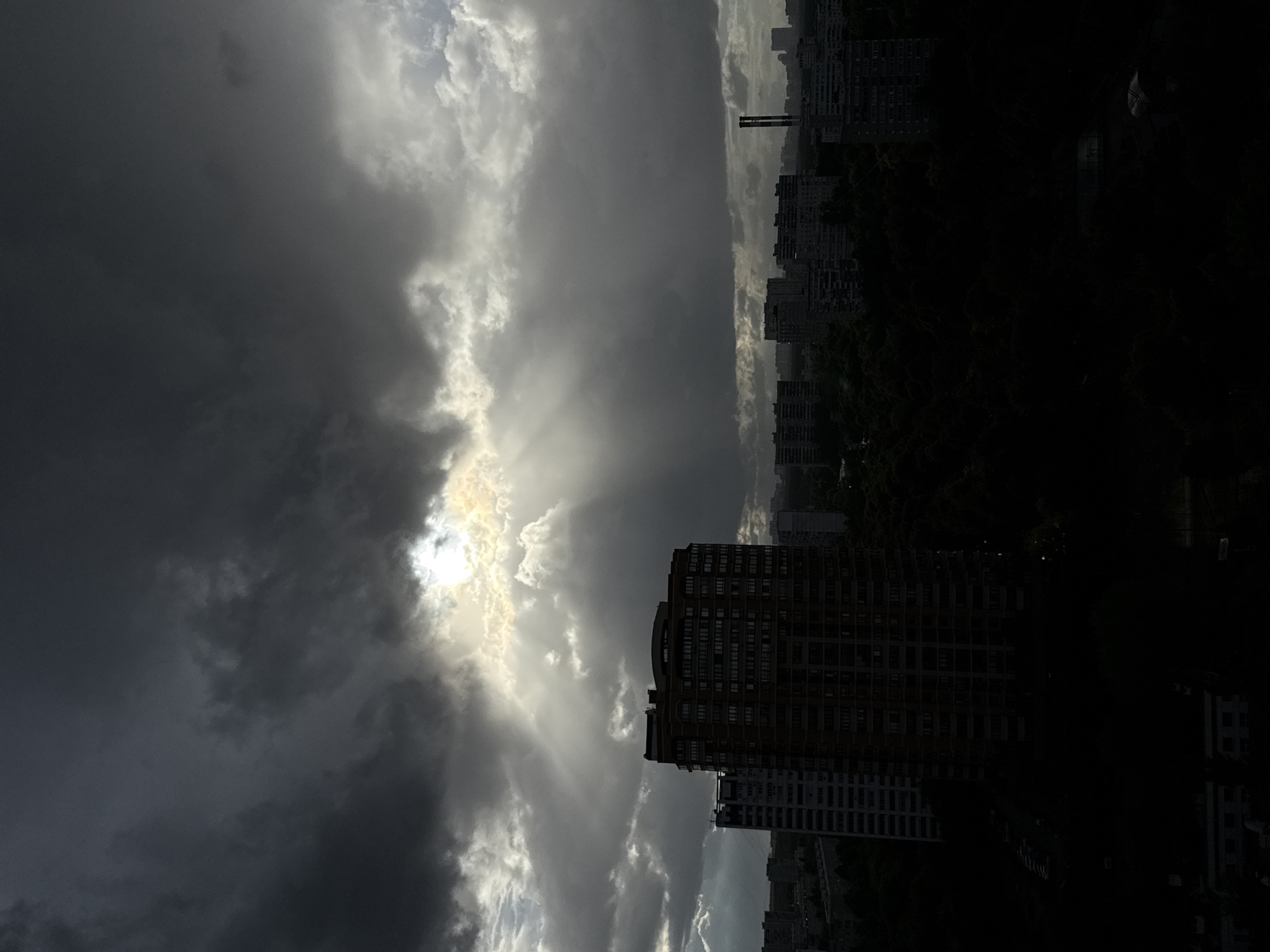 The image captures a dramatic sky over a dense urban landscape, likely Moscow, Russia, based on architectural styles. The sky is dominated by dark, heavy clouds, suggesting an approaching storm or recent rainfall. Sunlight breaks through a gap in the clouds, casting visible rays downwards, creating a dramatic, almost ethereal lighting effect. In the foreground, a tall, multi-story apartment building with a reddish-brown facade stands prominently, its windows reflecting the dim light. To its left, a taller, more slender building with a white facade is partially visible. The lower portion of the image is largely obscured by darkness, revealing silhouettes of more apartment buildings and dense trees, indicating a residential area with significant green spaces. The overall scene evokes a sense of foreboding yet also a striking natural beauty, as the light pierces the gloom.