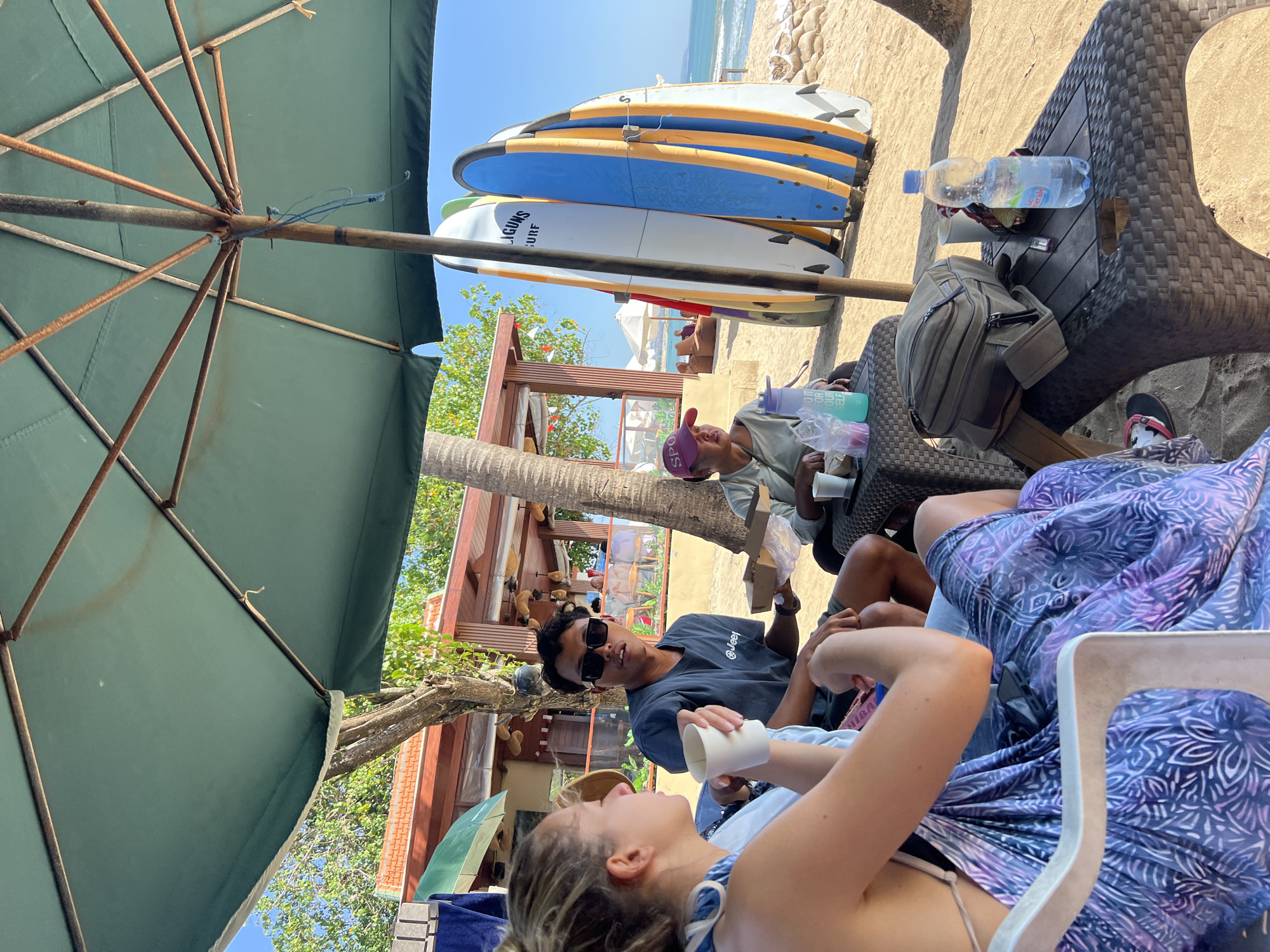 The image depicts a group of people sitting on chairs at a table on a sandy beach, likely in Kuta, Indonesia. A large green umbrella provides shade. In the foreground, a woman in a purple and blue sarong is seated, holding a white cup. Next to her, a man wearing sunglasses and a dark t-shirt is holding a small cardboard box. Another person, wearing a pink hat, is seated further down the table, holding a cup and a plastic bag. A table in front of them holds several items including cups, a plastic bag with food, a water bottle, and a backpack. In the background, several surfboards are propped upright, and a building with a red roof and outdoor seating is visible. Beyond the building, the ocean and a clear blue sky are present. The overall scene suggests a casual gathering on a beach, likely at a beachfront cafe or restaurant.