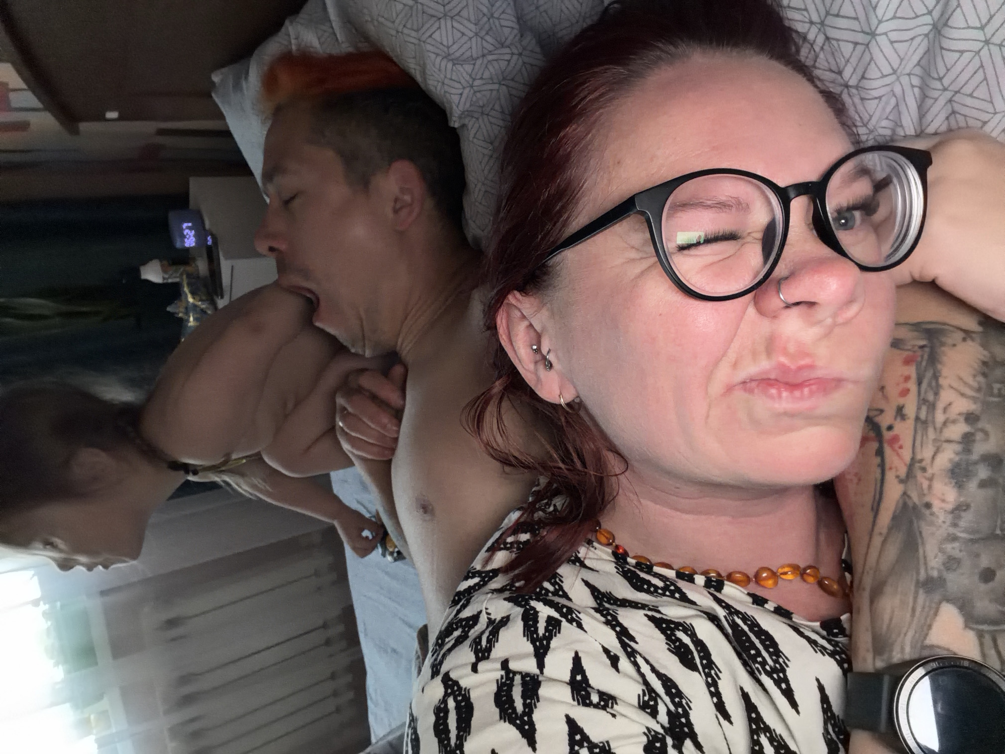The image shows two adults lying in bed. A person with red hair and round glasses is in the foreground, appearing to take a selfie. They are wearing a black and white patterned shirt and an amber necklace. Visible on their arm is a tattoo. In the background, another person with orange-tinged hair is yawning. A woman in the reflection is assisting the man from background by pulling out his arm. The bed has a white patterned duvet. A nightstand with an alarm clock and some other objects is visible near the bed. The location is presumed to be in Chaikovsky, Russia.