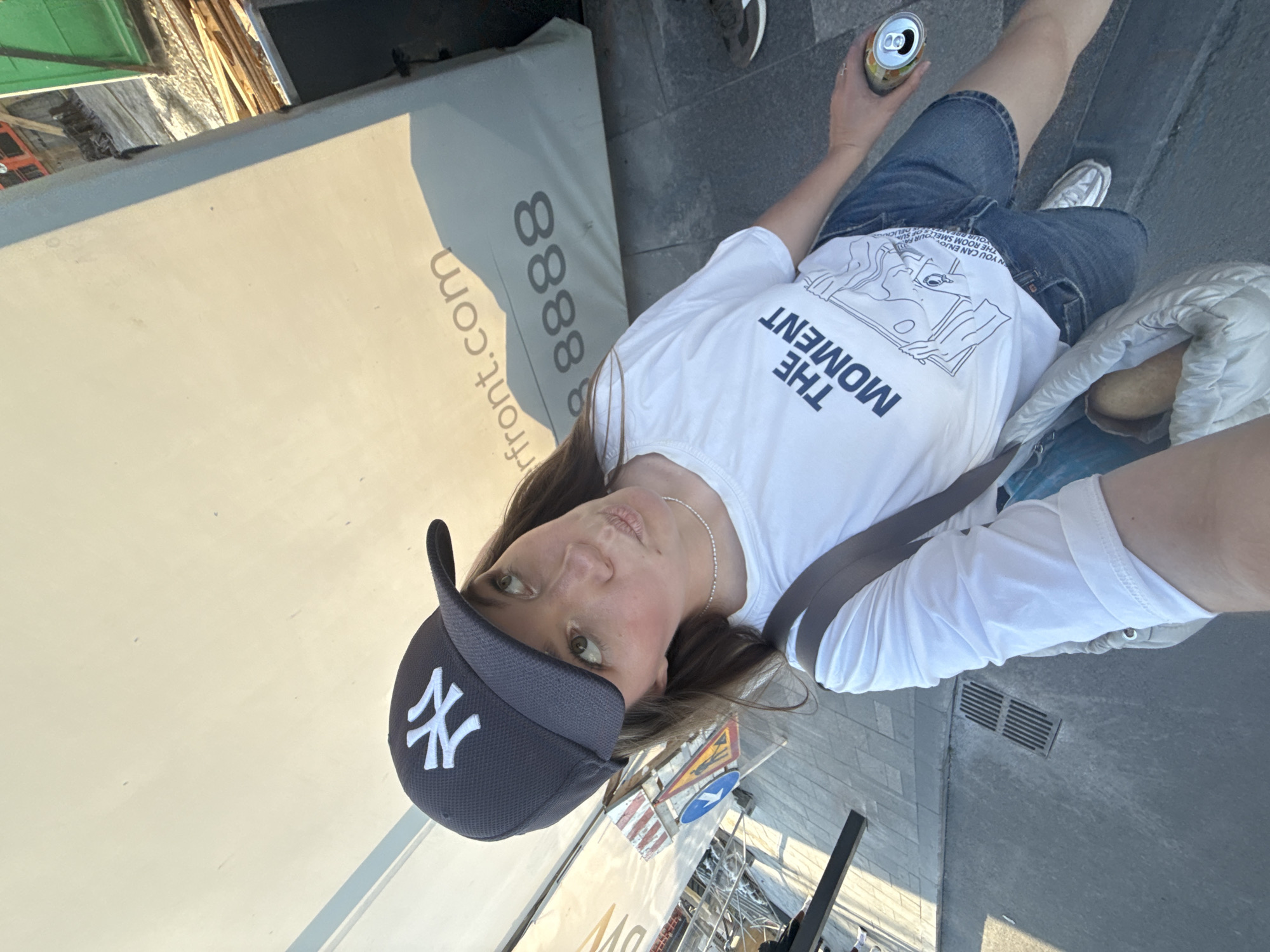 A woman with light brown hair is taking a selfie. She is wearing a navy blue New York Yankees baseball cap, a white t-shirt with the words "THE MOMENT" printed on it, and denim shorts. She is holding a can in her right hand and a white bag slung over her shoulder. She is standing on a city sidewalk. In the background, there is an advertisement sign with the words "front.com" and "88888". The location appears to be Belgrade, Serbia.