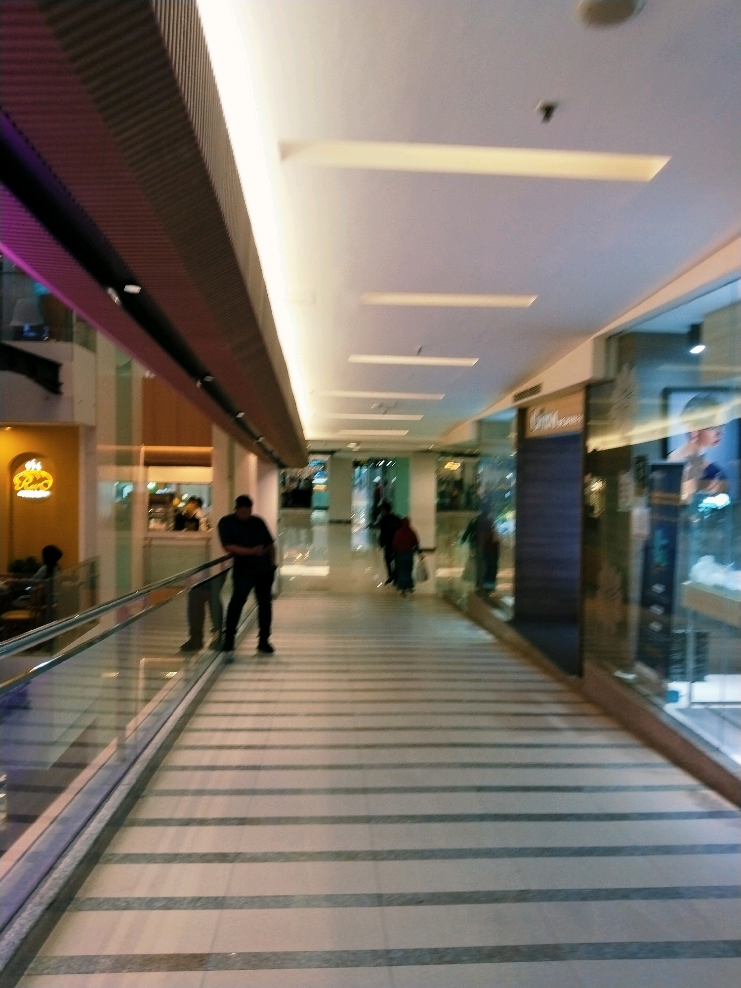 The image depicts a modern interior walkway, likely within a commercial establishment or shopping mall. The walkway features a checkered floor pattern with light and dark gray rectangular tiles, creating a sense of depth and perspective. On the left, a glass railing with a metallic base separates the walkway from a lower level, which appears to be a moving walkway or escalator. A lone figure, a man wearing a dark t-shirt and pants, stands leaning against the railing, facing away from the camera. His silhouette is clearly visible against the bright interior lighting.
Further down the walkway, several other individuals are visible, some walking and others standing, but their features are blurred due to motion or distance. To the right of the walkway, storefronts with large glass windows are apparent. One storefront prominently displays a sign with the word "Citra" and an advertisement featuring a human portrait. The overall environment is well-lit with a combination of ambient ceiling lights and strip lighting along the upper edges of the walkway.
The architecture suggests a contemporary design, with clean lines and materials like glass, metal, and polished stone. The purple lighting on the far left suggests the presence of decorative elements or potentially a specific zone within the establishment. Based on the stylistic cues and common commercial architecture in the region, this scene is consistent with an interior space in a modern shopping mall in Surabaya, Indonesia, such as Galaxy Mall or Tunjungan Plaza.