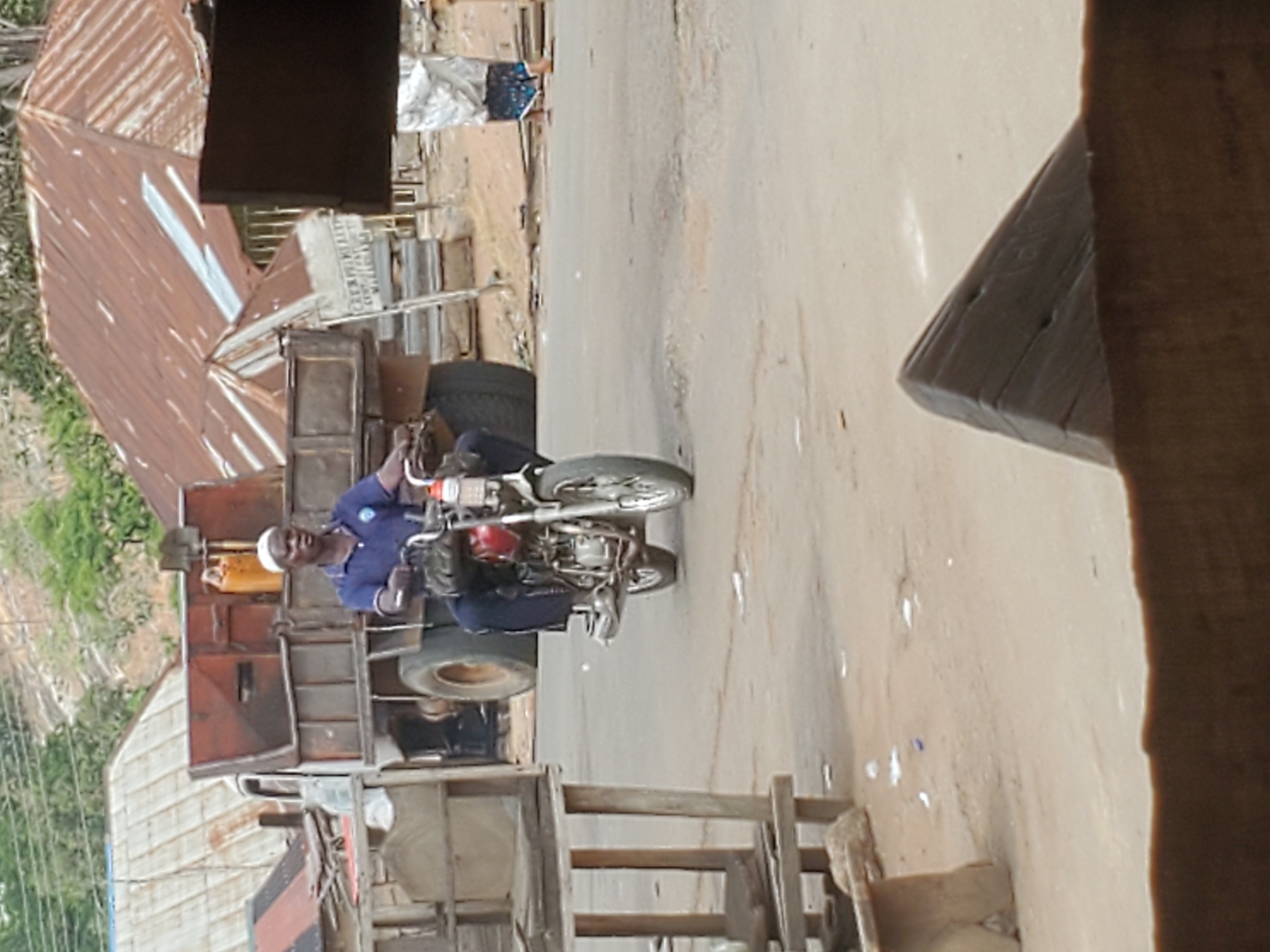 In Mararaba, Nigeria, a man wearing a white cap and a blue uniform sits on a red motorcycle on a paved street. The motorcycle is positioned in front of a truck with a rusty cargo area. Behind the truck and motorcycle, there are buildings, including one with a rusty corrugated iron roof. A wooden structure is present in the foreground. The background features green vegetation and hills.