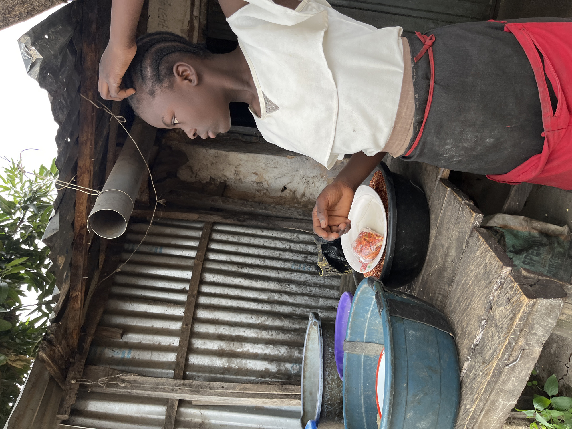 This image, captured in Jos, Nigeria, shows a young individual, likely female, engaged in a domestic task within a rustic, semi-outdoor setting. The subject, with dark skin and neatly braided hair, is depicted from the waist up, bending forward and looking down at several bowls. Her right hand rests near her head, while her left hand is positioned over one of the bowls. She wears a white, layered top over a dark grey or black garment, with a red tied apron or piece of cloth around her waist.
In front of her, on a wooden surface, is a black basin filled with numerous small, reddish-brown grains, possibly sorghum or millet. A white bowl sits atop these grains, containing a clear plastic bag with small, dried, reddish-orange items, which could be spices or dried food. To the left, a blue plastic basin and a purple basin are visible, along with another white/grey basin.
The background reveals a simple structure made of corrugated metal sheets and rough wooden planks, suggesting an unsophisticated, possibly improvised, shelter or workspace. A grey metal pipe is visible in the upper left, attached to the structure. Green foliage from nearby trees or plants can be seen in the upper left. The lighting is bright and diffused, indicating daytime, possibly under an overcast sky or indirect sunlight. The overall scene conveys a sense of daily life and routine in a modest environment. There is no visible text in the image.