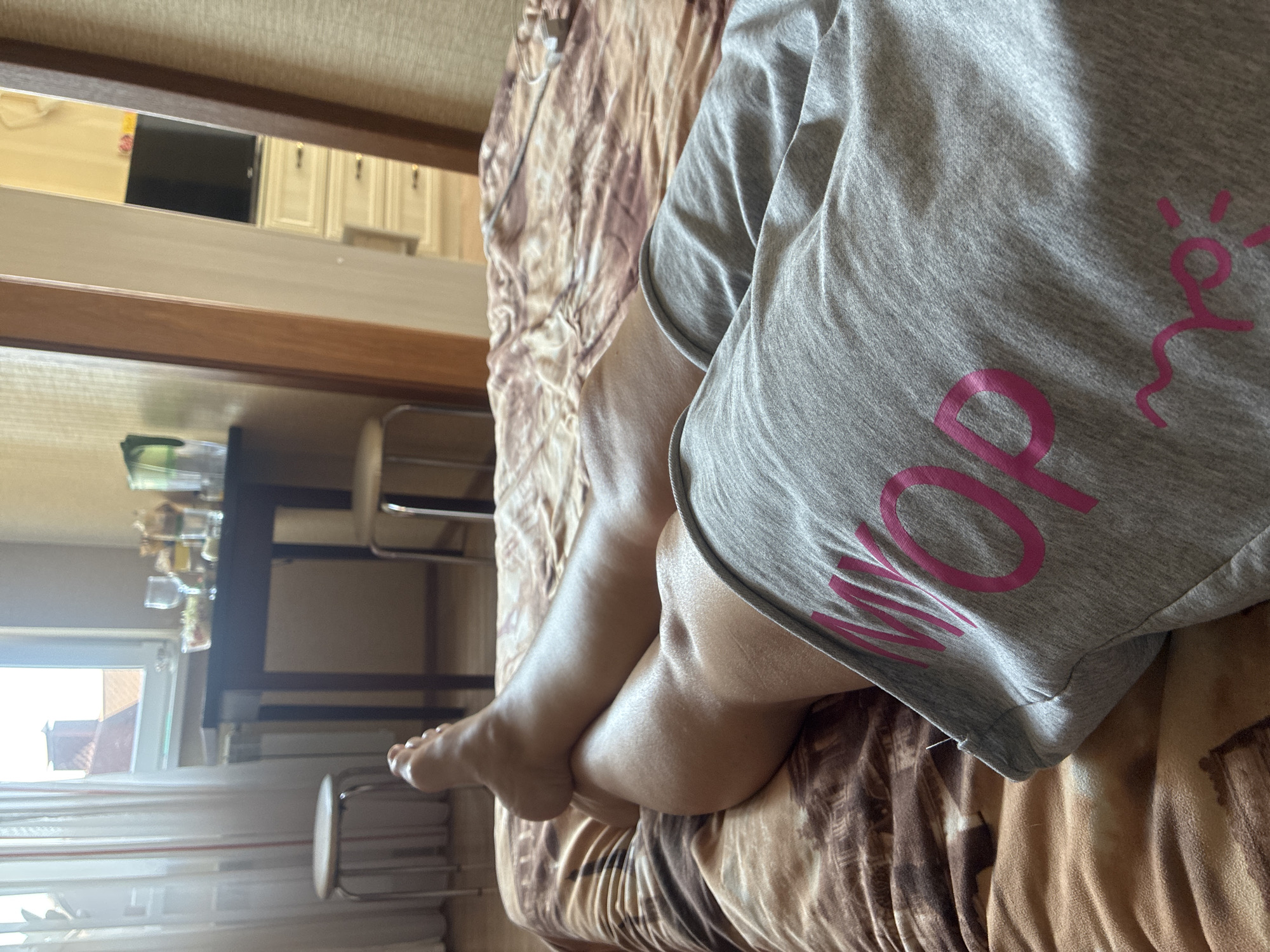 The image depicts a person lying on a bed covered with a brown and beige patterned duvet. The person is wearing grey shorts with pink lettering that reads "MOP". The focus of the image is on the person's legs. In the background, a dark wooden table with glasses and food items is visible. To the left of the table, a white stool and a window with sheer white curtains are present, through which a rooftop and sky can be seen. To the right, a doorway leads to another room, where a television and white cabinets are visible. The scene appears to be indoors, possibly in a bedroom or living area. There is no indication of the location being in Sochi, Russia.