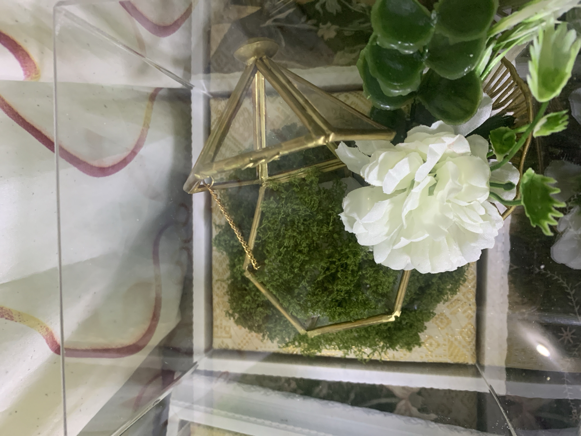 The image shows a close-up of a decorative arrangement inside a glass container. The arrangement features a hexagonal geometric terrarium made of brass-colored metal and glass, filled with green moss. A white artificial flower with layered petals is positioned in front of the terrarium. To the right of the flower are several round, glossy green leaves. The background is out of focus but appears to be a patterned fabric. The scene is indoors, likely a display or gift item. There are no people or animals visible, and no specific location in Kuala Pembuang, Indonesia can be identified from the image.