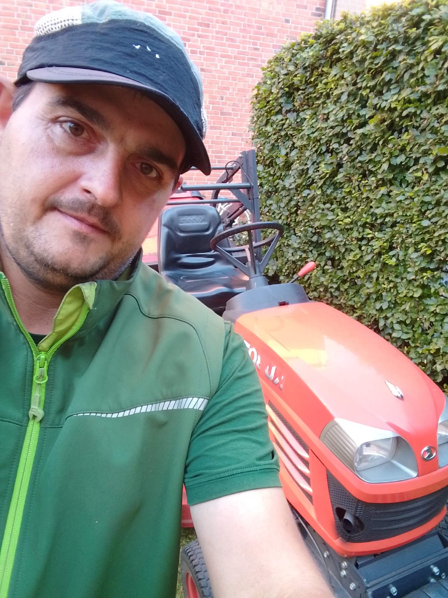 A man wearing a green jacket and a baseball cap is taking a selfie next to a red tractor. The man is positioned in the foreground, with the tractor and a hedge in the background. The tractor has a black seat and steering wheel, and its front end is visible, featuring headlights and a grille. The hedge is dense and green, and a brick wall is visible behind it. The overall scene appears to be an outdoor setting, possibly a garden or yard.