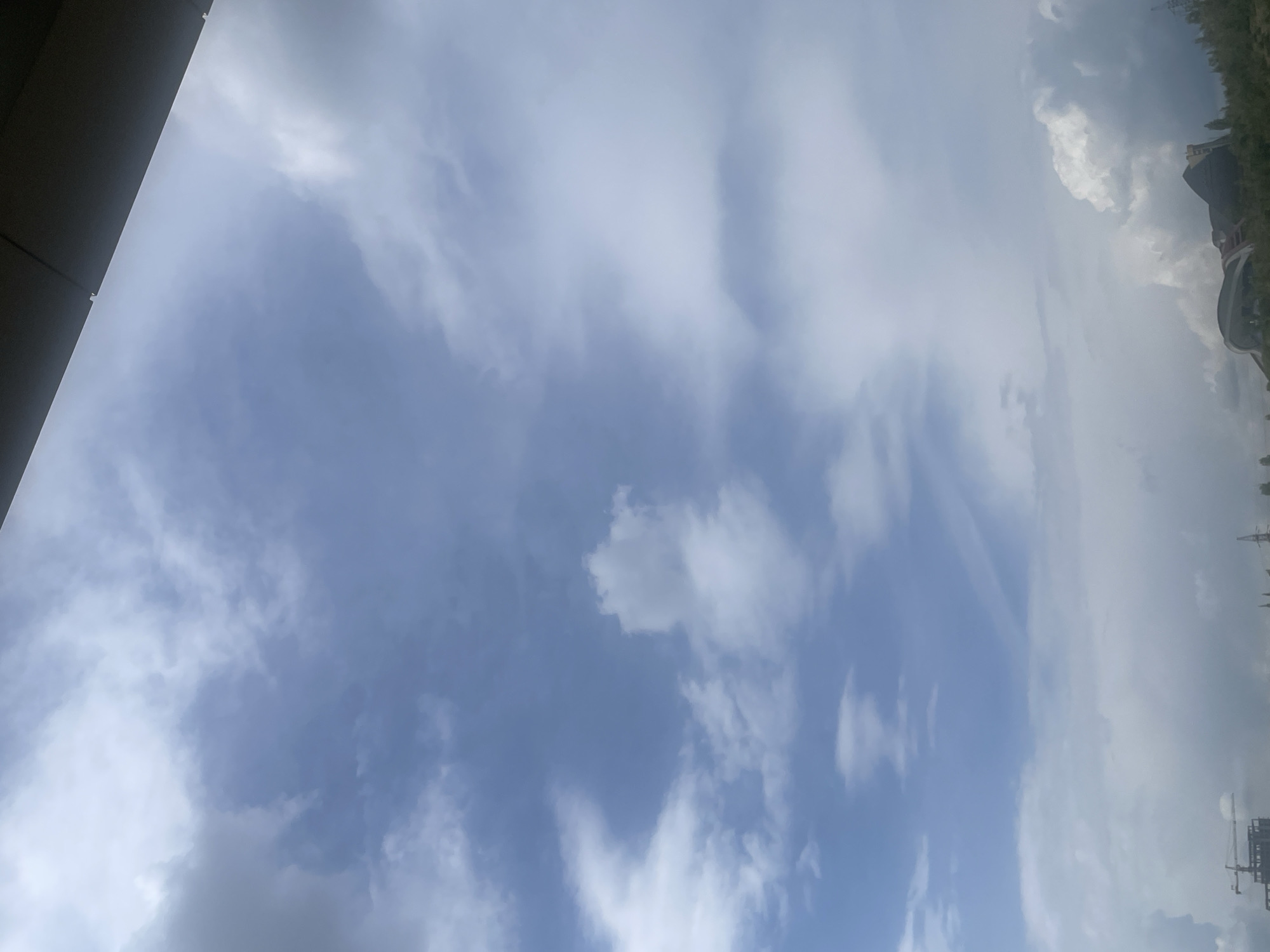 The image captures a partially cloudy sky over Yerevan, Armenia. The view is angled upwards, showing a building edge on the left side of the frame. The sky consists of a mix of light blue patches and white, wispy clouds. Part of the Yerevan skyline is visible on the right side, with indistinct buildings and power lines. The photograph seems to have been taken from a low angle, potentially within a building or near a tall structure.