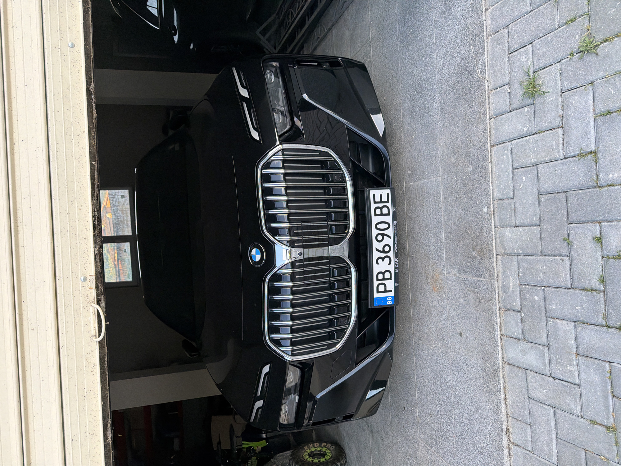 The image shows the front of a black BMW 7 series car parked partly in a garage. The license plate reads "PB 3690 BE" with the country code "BG," indicating Bulgaria. The car is partially situated on a gray paved surface with some vegetation growing in the gaps between the pavers. The garage has a rolling door with a handle. The license plate indicates a possible origin in the Blagoevgrad Province of Bulgaria. The exact location cannot be determined to be within the city of Asenovgrad specifically as it is not indicated on the license plate. A green and black object is partially visible on the lower left.