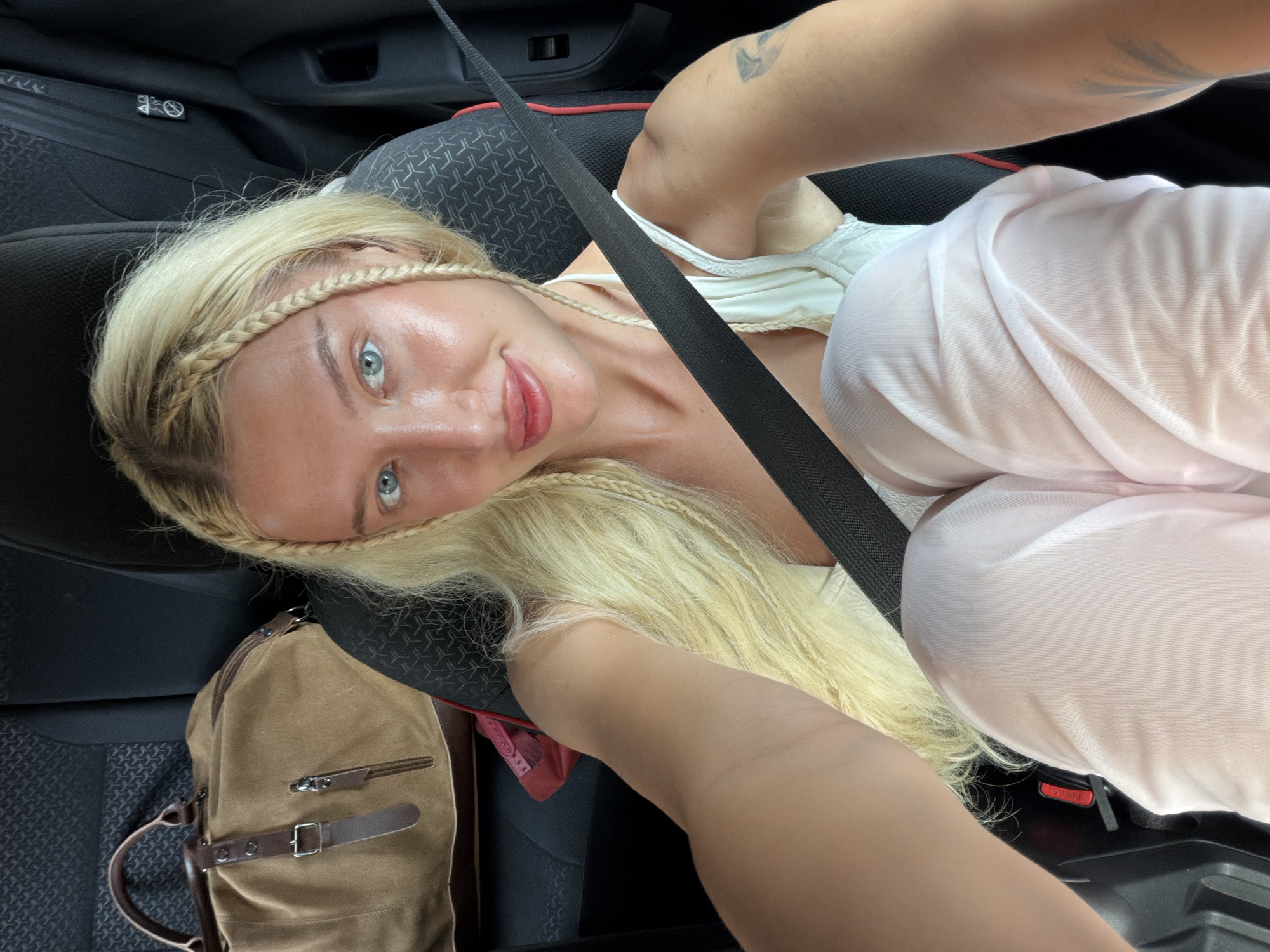 A young blonde woman with blue eyes and blonde braided hair is taking a selfie in the backseat of a car. She is wearing a white tank top and light pink pants. A large brown duffel bag is on the seat next to her. A seatbelt is across her chest. The interior of the car is visible, including the seats and the car door.