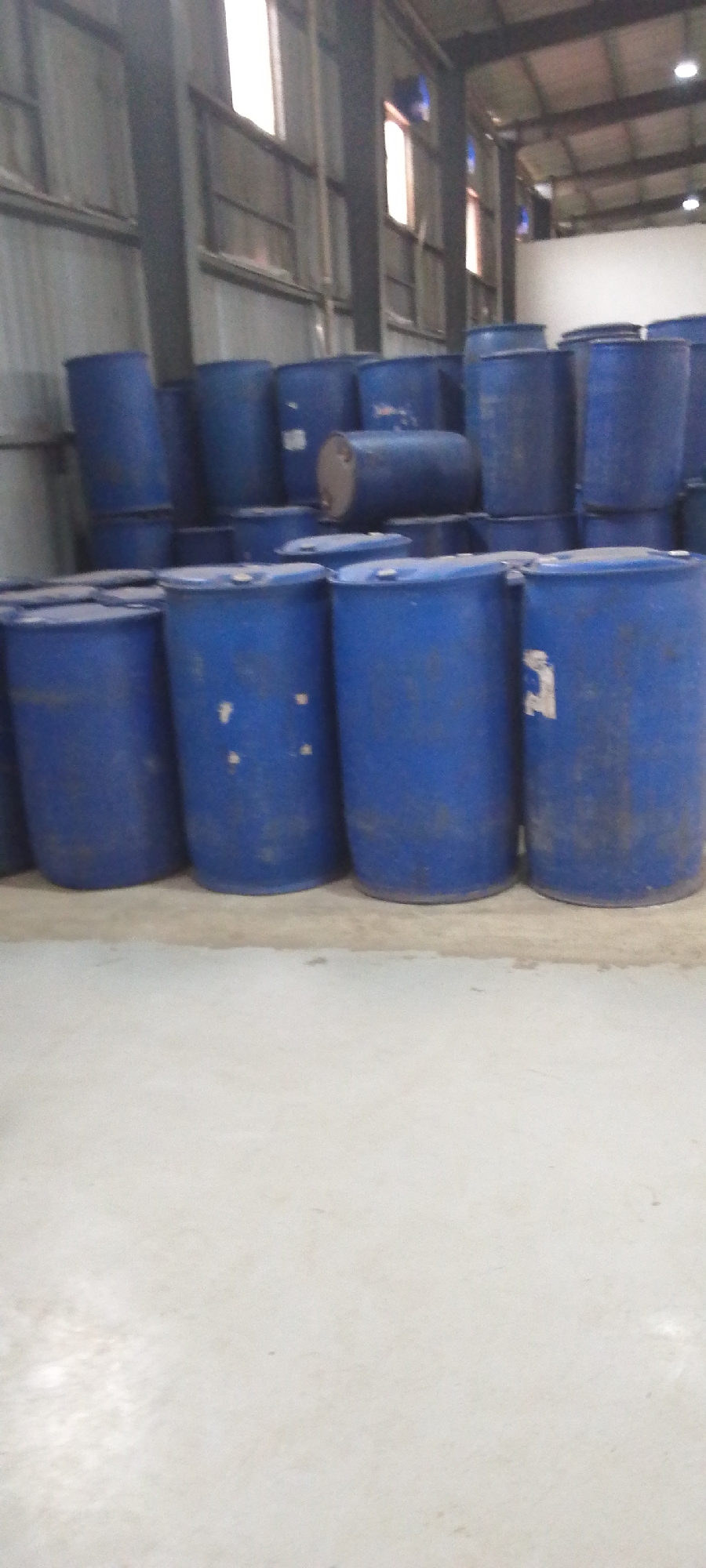 The image depicts a large industrial warehouse in Unknown, Unknown filled with numerous blue plastic barrels. The barrels are stacked in rows, some sitting horizontally atop others. The warehouse appears to have a high ceiling with metal support beams and several windows allowing natural light to enter. The floor is made of concrete. No people are visible. The general environment suggests a storage facility for industrial materials.