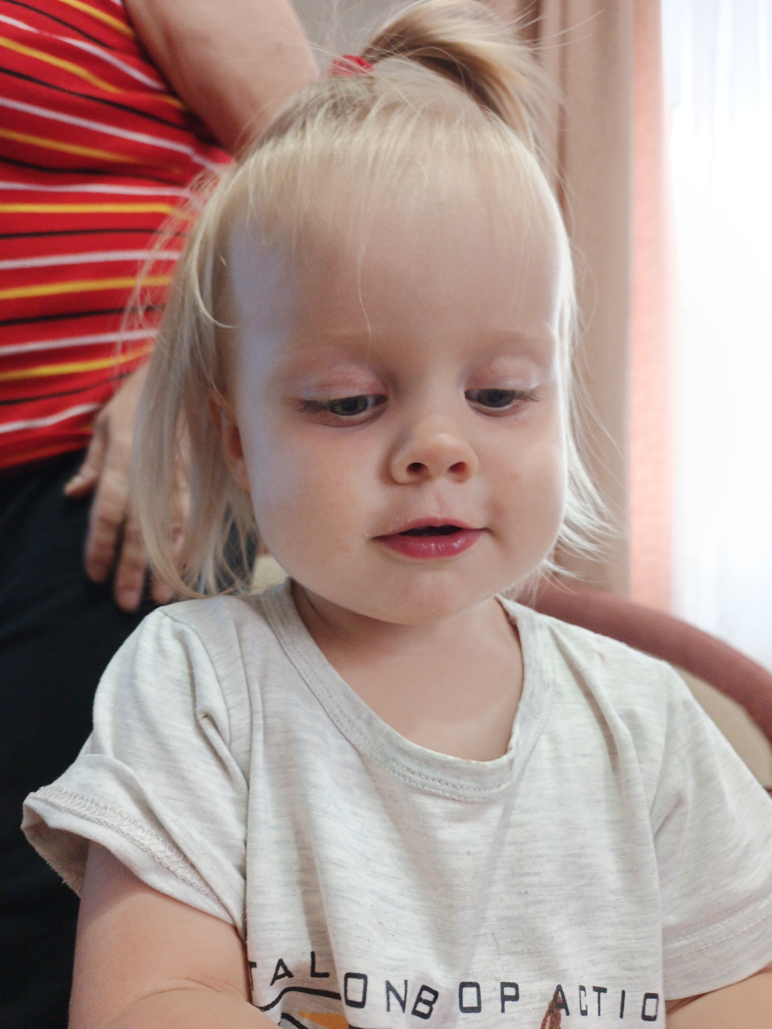 A close-up, vertical, medium shot captures a young Caucasian child from the chest up, seated and looking down. The child has blonde hair pulled into a high ponytail and is wearing a light gray t-shirt. The t-shirt has a graphic design on the front, partially visible, with black letters and some yellow and orange accents. The background is out of focus and includes a person wearing a red and yellow striped shirt to the left, and a curtain or fabric in a peachy hue to the right, suggesting an indoor setting. The lighting appears to be natural, coming from the right. There is no information about the specific location within Medvedyovka, Russia, other than the city name itself.