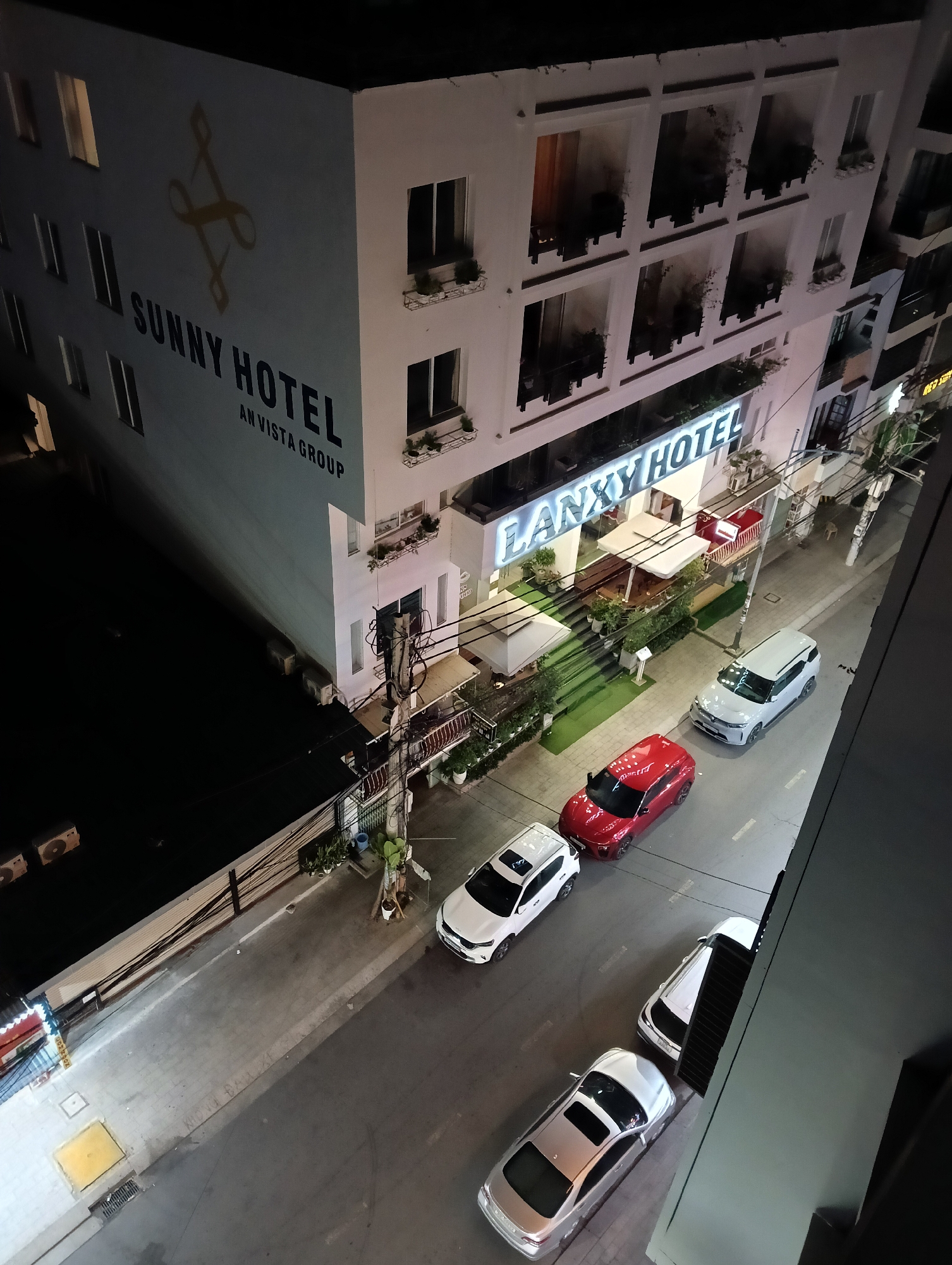 An aerial view at night shows a street in Nha Trang Ward, Vietnam, with two hotels, "Sunny Hotel" and "Lanxy Hotel." The street contains four parked cars: silver, red, and two white. A utility pole with multiple wires stands on the sidewalk between the two hotels. Two people are on the sidewalk by the utility pole.