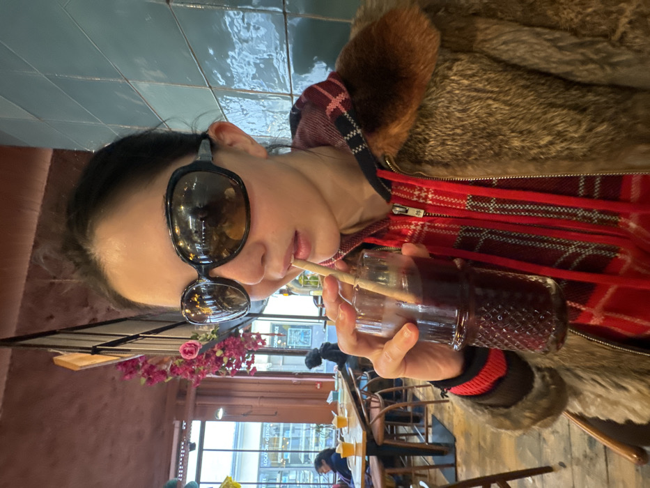 A person, appearing female, is seated indoors in what resembles a cafe or restaurant, captured from the upper torso. She wears large, dark, oval-framed sunglasses, a red plaid zippered jacket, and a brown fur-collared coat. She holds a textured glass containing a dark reddish-purple beverage and drinks from it via a light-colored straw inserted between her lips.
The interior environment features a wall of glossy light teal square tiles to the right. To the left, a large window overlooks an urban street with buildings and displays French signage reading "LOCATION - GESTION" and "COMMERCE," identifying the setting as Paris, France. Pink floral decorations hang from a reddish-brown ceiling element, and out-of-focus tables, chairs, and other patrons are visible in the background.