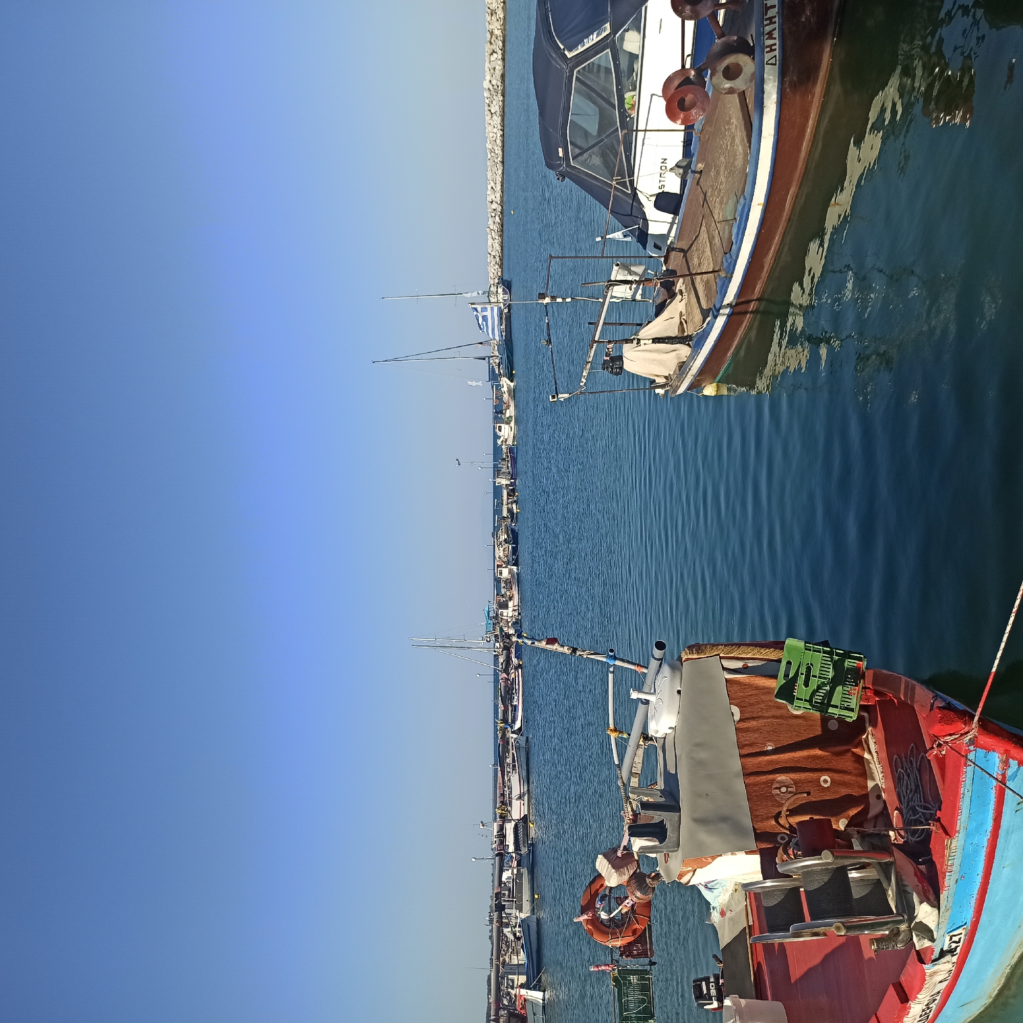 In the city of Limani Litochorou, Greece, a bright, sunny day unfolds with a clear blue sky overhead. The scene is a harbor with calm, deep blue water reflecting the bright daylight. Two fishing boats are prominently featured in the foreground, both docked and appearing stationary. The boat on the left is red and blue with various fishing equipment visible, including a life preserver and a green plastic crate. The boat on the right is a darker brown and white vessel, with a covered cabin and some mooring lines. In the background, a line of more boats, including sailboats and smaller fishing boats, are moored along a breakwater constructed of large rocks. The distant horizon shows a faint outline of land under the expansive sky. There are no people visible in the image.
