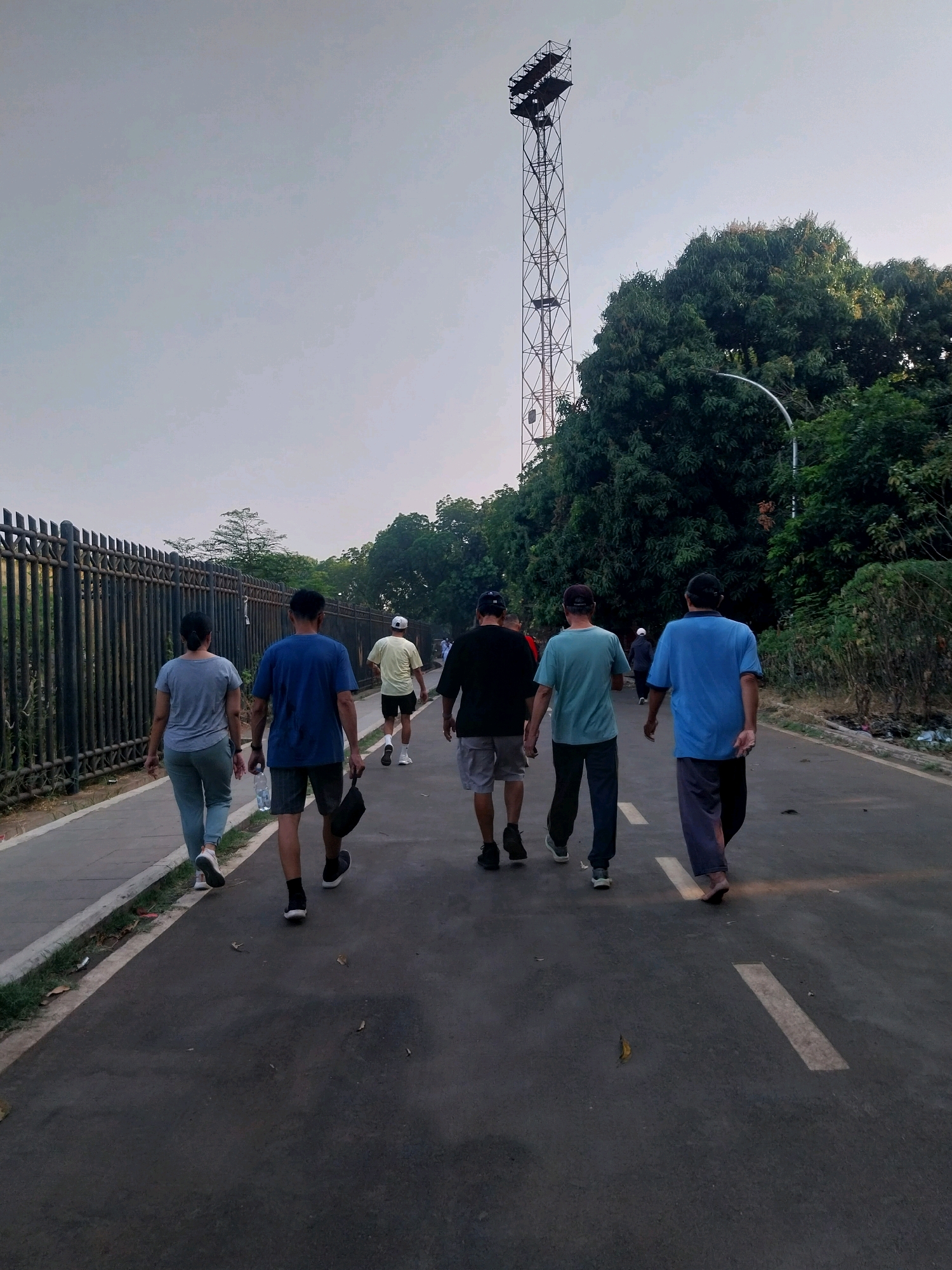 A group of six individuals, viewed from behind, are walking away from the camera on a paved path. The path is bordered by a tall, dark metal fence on the left and lush green trees and foliage on the right. A tall, metal tower, possibly a communication or lighting tower, stands against a pale sky on the right side of the image, behind the trees. The individuals are dressed in casual attire, including t-shirts, shorts, and long pants. One person is carrying a black bag. The scene appears to be an outdoor recreational area, likely a park or promenade, in the city of Cirebon, Indonesia. The lighting suggests it is either early morning or late afternoon.