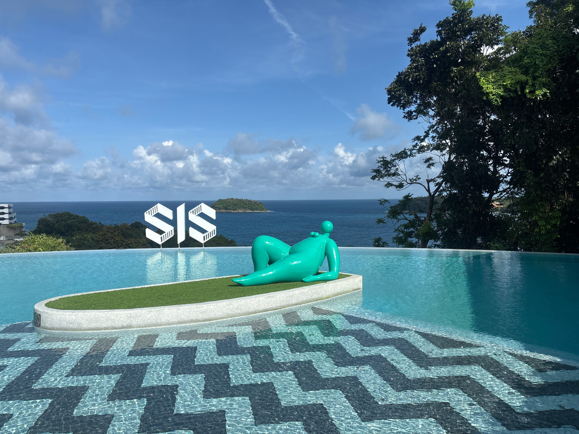 The image shows an infinity pool with a teal statue of a reclining figure on a rectangular platform covered with artificial grass. The pool's interior features a black and blue zigzag tile pattern. In the background, across the ocean, a small green island is visible. Above the island, a white logo consisting of three abstract shapes that resemble stylized 'S' letters is displayed on a pole. The sky is blue with scattered white clouds, and lush green trees are present on the right side of the frame. The location is the city of Karon, Thailand.