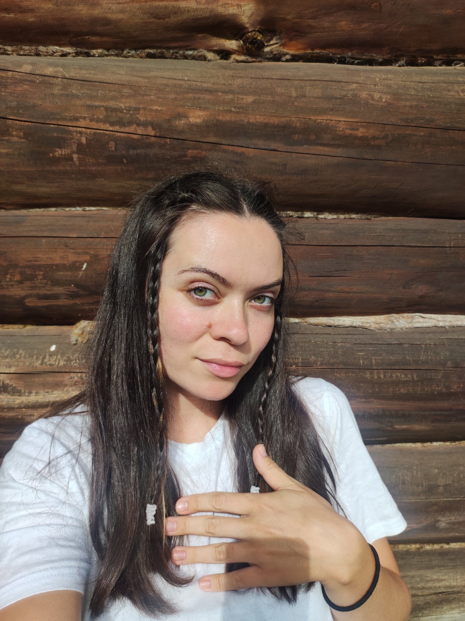 A female subject is depicted from the chest up, positioned in front of a rustic log wall in Фоки, Russia. She has long, dark brown hair, styled with multiple thin braids framing her face, some adorned with light-colored wraps. Her green eyes are open, and she looks directly at the viewer with a neutral expression. A small, dark mole is visible on her right cheek (viewer's left). She wears a plain white short-sleeved t-shirt with a crew neck. Her right hand (viewer's left) is placed over her left chest, palm down, with visible nail beds. A black elastic band is present on her left wrist (viewer's right). The background consists of horizontally stacked, dark brown logs, likely from a cabin or similar wooden structure. Gaps between the logs are filled with a lighter, fibrous chinking material. The scene is illuminated by strong natural light, casting shadows on the subject's left side of the face (viewer's right) and her right hand.