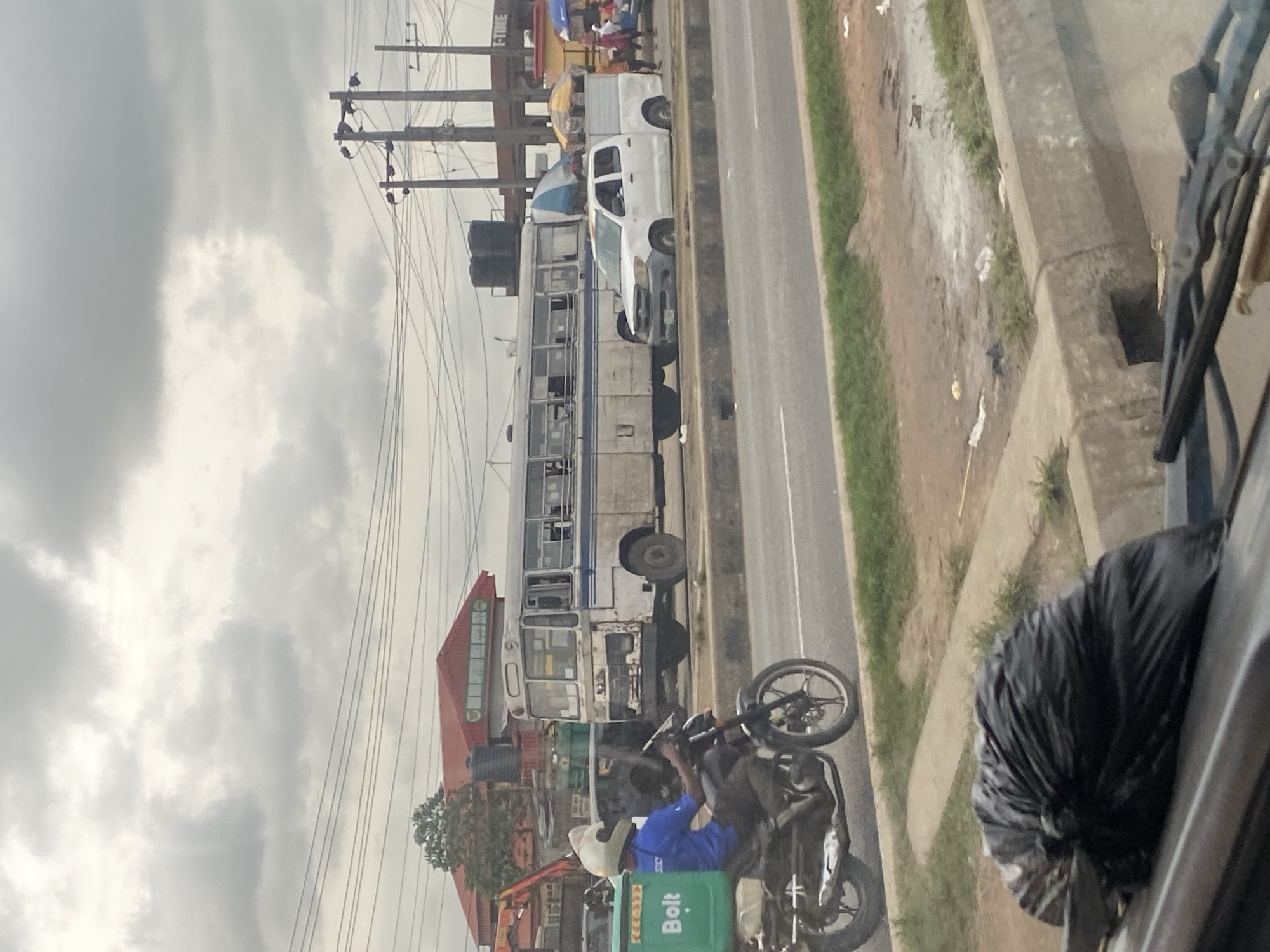 This is a medium shot, eye-level, vertical street scene in Ikeja, Nigeria, under an overcast sky. In the foreground, a black trash bag rests on the hood of a vehicle, with windshield wipers visible. The middle ground shows a road with a concrete divider and grass. On the road, a person wearing a blue shirt and white helmet is riding a motorcycle with a green "Bolt" delivery box on the back. To the right of the motorcycle, a white pickup truck is parked, and further to the right, a white and blue bus is stopped. People are visible near the bus and pickup truck. In the background, a red-roofed building with large windows is visible, along with utility poles and wires, and other structures with signs. The environment appears somewhat cluttered with debris on the side of the road.