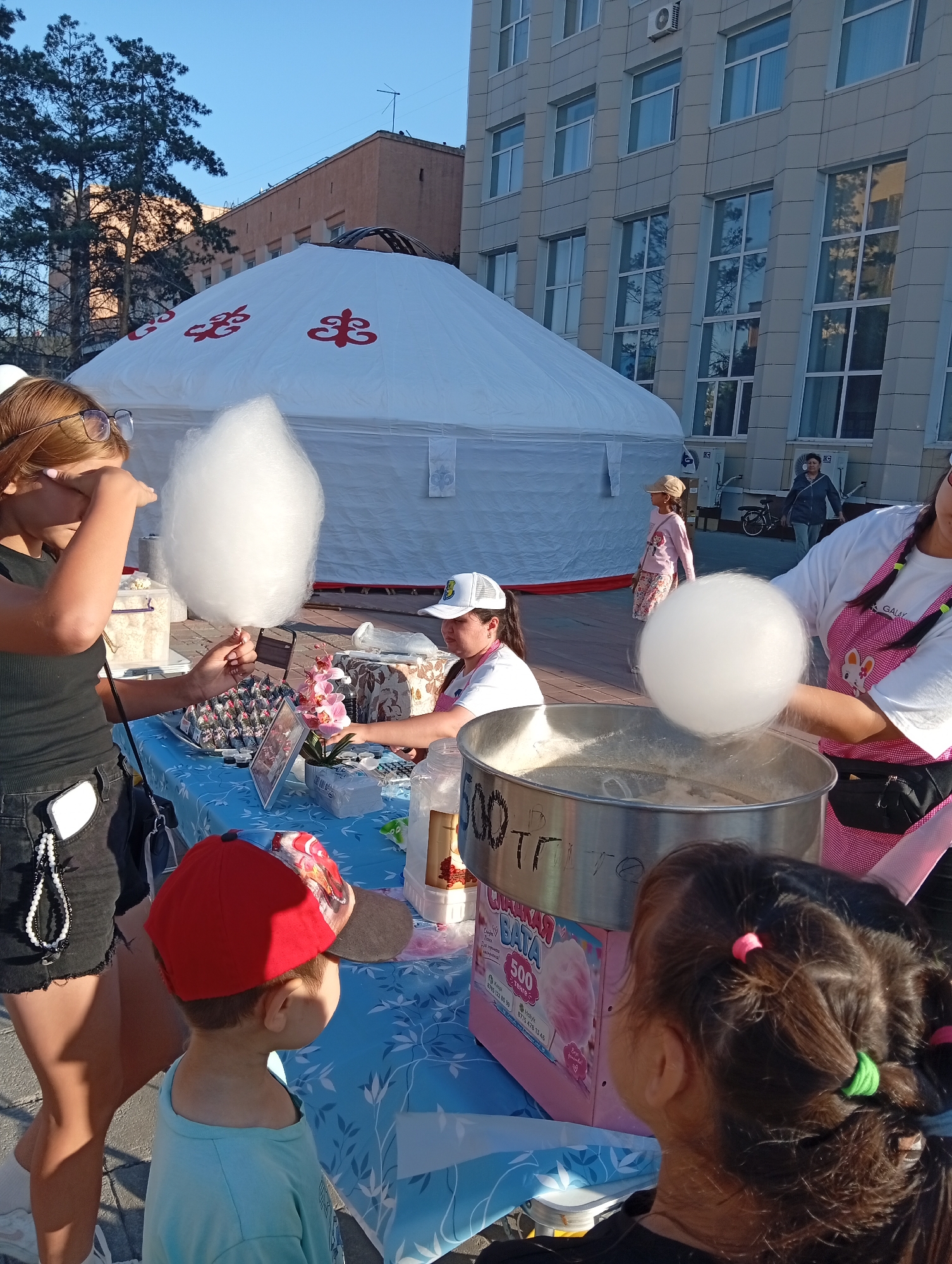The image captures an outdoor scene in Kostanay, Kazakhstan, likely on a sunny day. In the foreground, a girl with reddish-brown hair and glasses holds a large, fluffy piece of cotton candy. She appears to be shielding her eyes with her other hand. Beside her, a young boy in a red baseball cap and a light blue shirt looks to his right. A vendor, wearing a white cap and a pink apron adorned with cartoon rabbits, is operating a cotton candy machine. The machine is silver and pink, with a label that reads "СЛАДКАЯ ВАТА 500" (Cotton Candy 500). A pink cotton candy cloud is visible emerging from the machine. The table in front of the vendor is covered with a blue tablecloth with a white floral pattern. On the table are several small packages, possibly snacks, and a small pink flower arrangement. In the background, a large, white yurt with red decorative symbols is prominently displayed. Behind the yurt, a modern, light-colored building with many windows dominates the skyline. Further in the background, other people are visible, including a person walking and another on a bicycle. Trees with green foliage are also present. The ground appears to be paved. The overall atmosphere suggests a public event or festival.