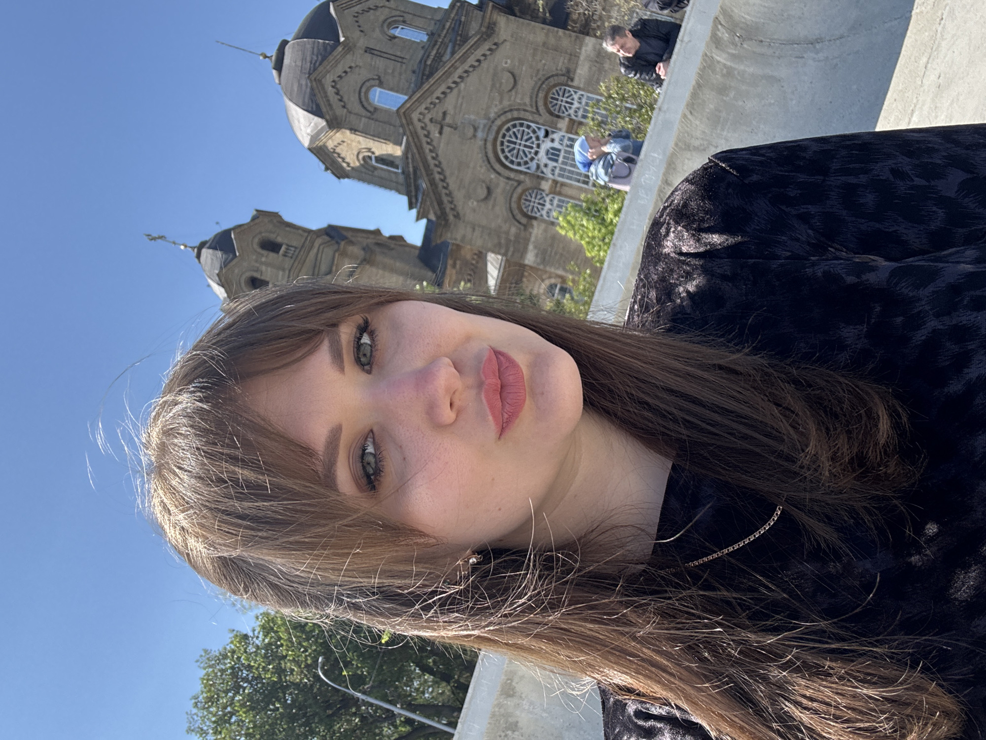 A young woman is posing for a selfie, outdoors, with a church in the background. The woman has long brown hair with bangs, fair skin, green eyes, and is wearing a dark patterned garment. She is wearing a delicate necklace. Behind her is a large, ornately designed church with twin domes and arched windows. There is a concrete structure in the foreground, possibly a wall or railing. In the far background, there are other people and trees. The sky is clear and blue. This church is likely located in the city of Yevpatoriya, Ukraine.