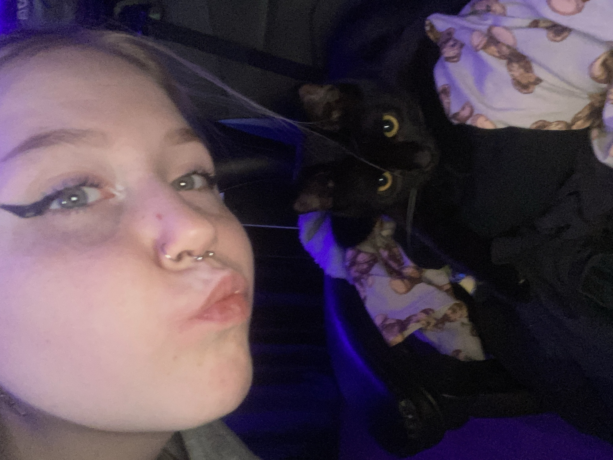 The image shows a selfie of a young woman with fair skin and visible eyeliner. She has a nose piercing and is making a kissing face towards the camera. Behind her, partially obscured by dark clothing, is a black cat with yellow eyes. A blanket with a teddy bear pattern is visible in the background. The location is not discernible from the image, and there's no evidence to suggest it is in Izhevsk, Russia.