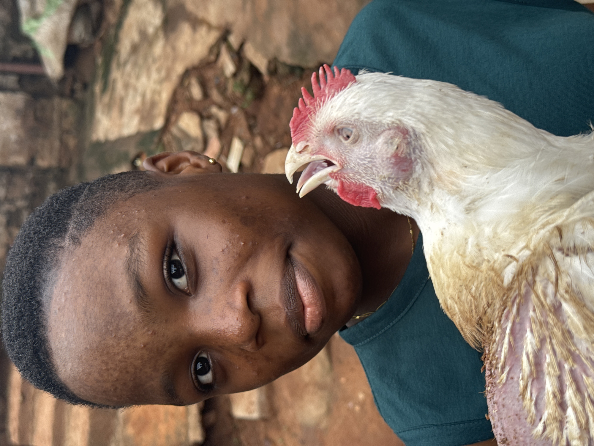 A person with dark skin and short black hair looks at the camera while holding a white chicken in their arms. The chicken has a red comb and appears to be squawking with its beak open. The person is wearing a teal shirt, and the background appears to be a stone or brick wall, possibly outdoors. The location is described as Onitsha, Nigeria.
