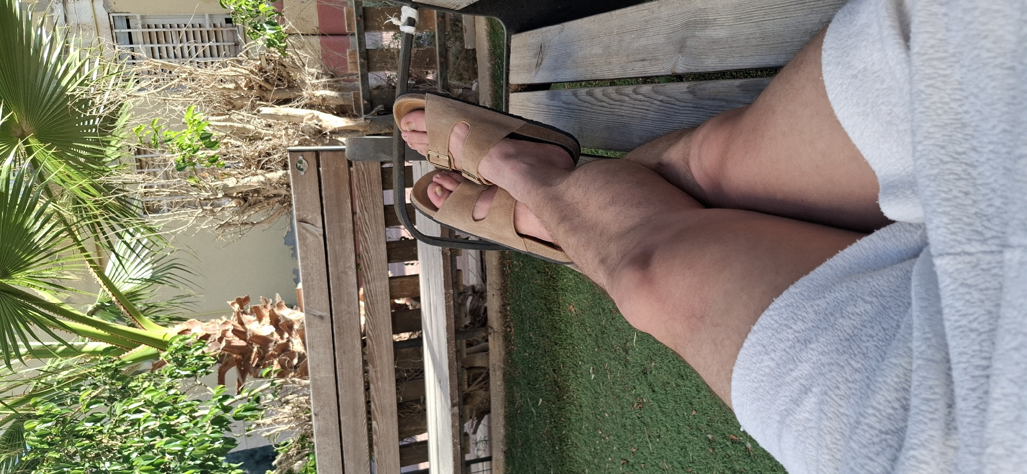 The media file shows a close-up of a person's legs and feet, wearing tan Birkenstock sandals and light-colored shorts or robe. The person is sitting on a wooden bench, with their legs crossed. Behind them, there is a green grassy area and some dry palm fronds and branches. A building with a barred window is visible in the background on the left. The lighting suggests it is daytime, likely sunny given the shadows. The overall scene appears to be outdoors, possibly in a backyard or patio area. There is no visible text in the image. The context provided indicates the location is Yerocham, Israel.