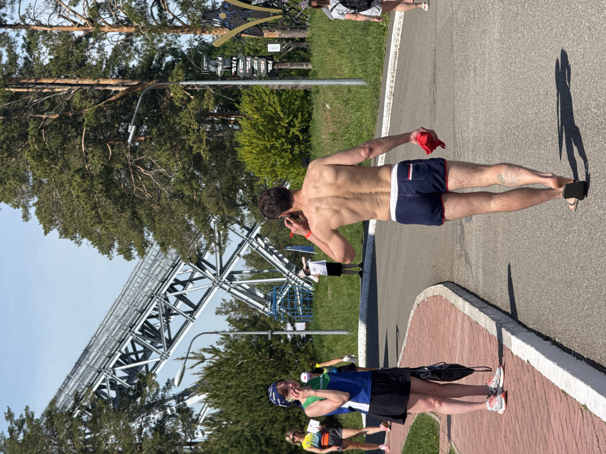 The image depicts a scene in what appears to be a park or recreation area in the city of Unknown, Unknown. Several individuals are present, engaged in various activities.
In the foreground, a woman in athletic attire (blue shirt and black shorts) is walking along a paved pathway. Nearby, a group of people in running gear are seen in the distance.
A shirtless man in dark blue swim trunks with a white stripe is also walking along the pathway. He is holding a red cloth or garment and appears to be talking on the phone.
In the background, there is a large metal structure, possibly a roller coaster or other amusement park ride. The sky is clear and blue. There is also vegetation, including trees and shrubs, visible in the scene. A signpost with various directions is visible in the middle of the frame. The pathway appears to be bordered by grass and a brick or stone edge.