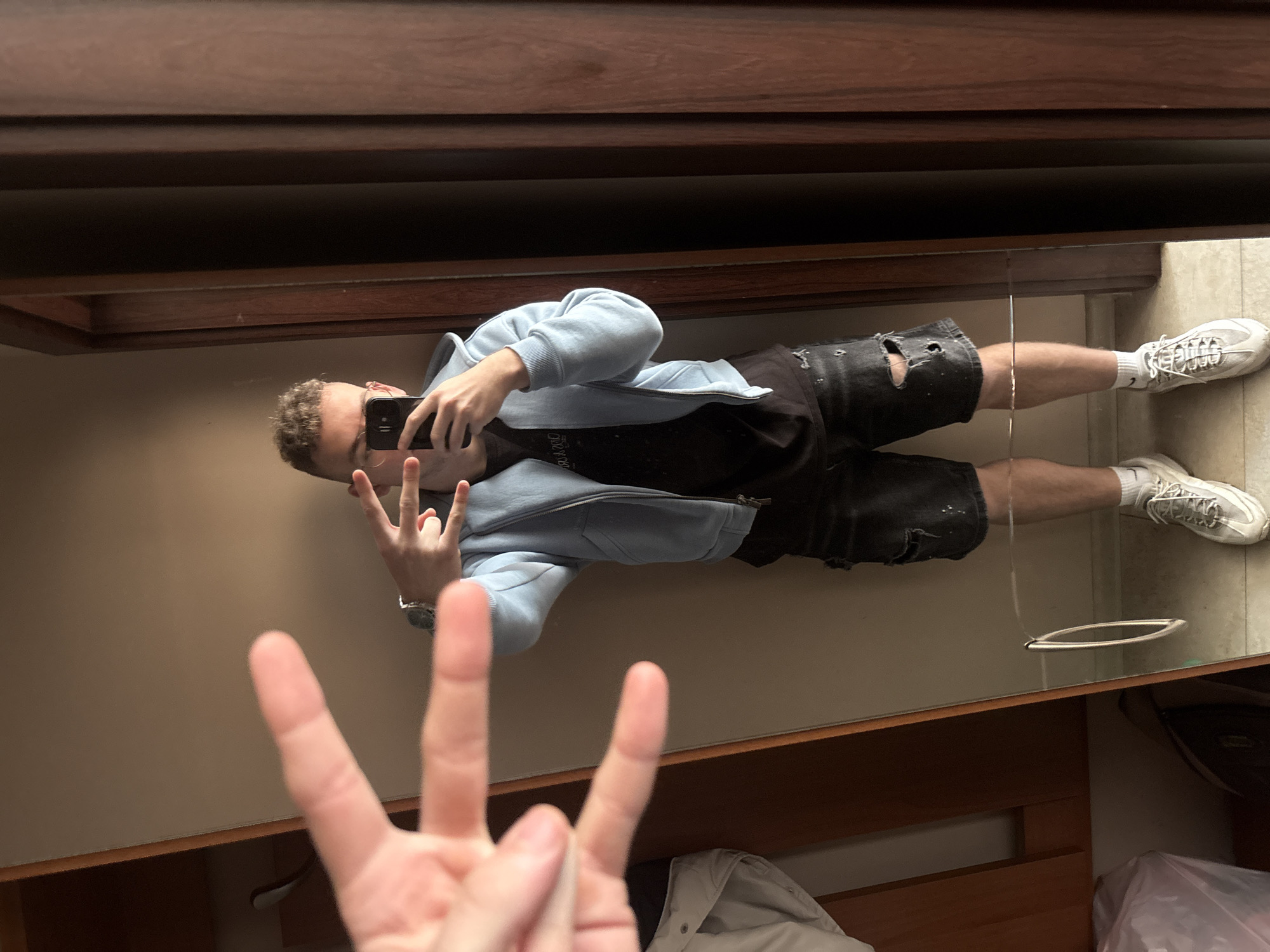 A fair-skinned individual, likely male, is reflected in a mirror within a room. The individual is wearing a light blue zip-up hoodie, a black t-shirt, ripped black shorts, white socks, and white sneakers. He is holding a black smartphone in his left hand and appears to be taking a selfie, making a hand gesture with the other hand. The reflection includes another hand making a different gesture and a glimpse of a white jacket or garment in the bottom of the frame. The background suggests the room has dark wooden trim. The location cannot be determined.