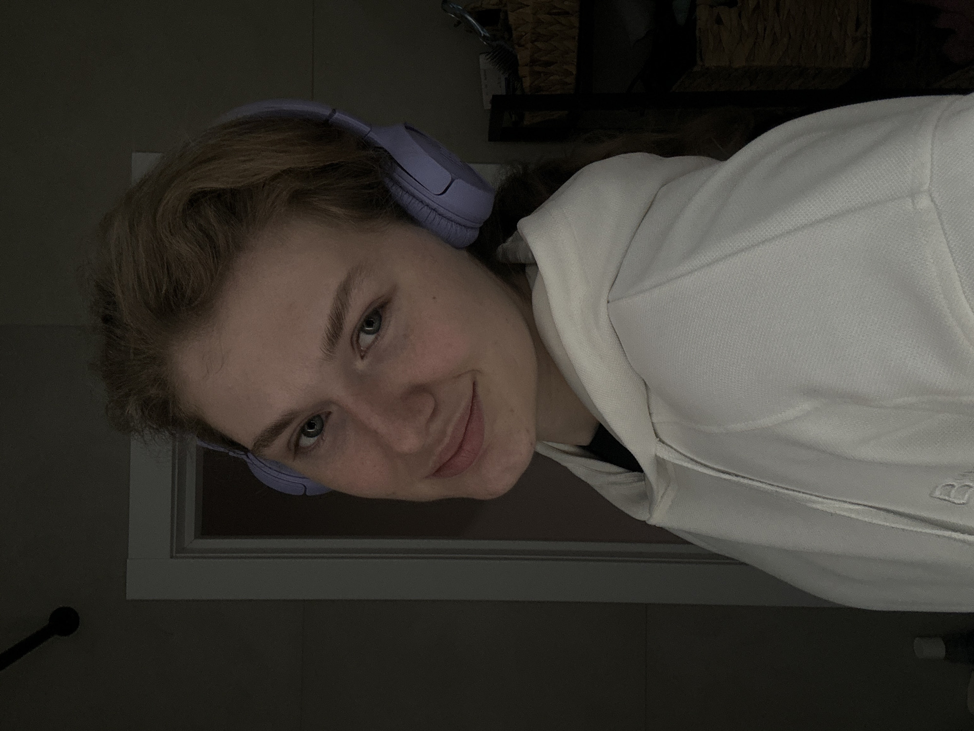 A young woman with light skin, wearing light purple headphones and a white hooded sweatshirt, is the main subject of the photograph. The scene appears to be indoors, with a window frame visible behind her head. The lighting is dim, making the background largely dark. Other objects visible in the background include a comb or brush and a wicker basket. The location cannot be determined based on the image.