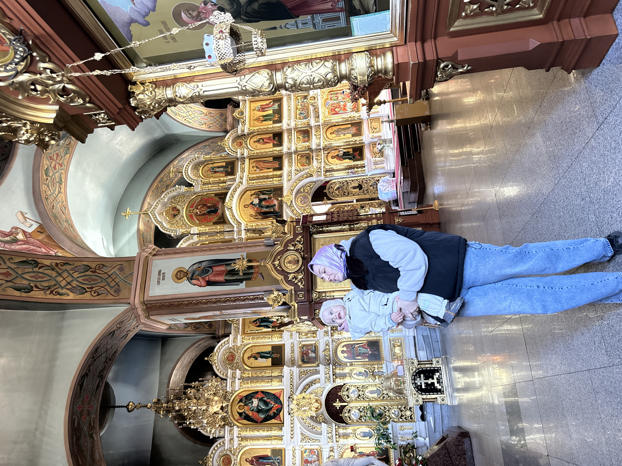Here is a detailed description of the image:
**Scene:** The image is taken inside an ornate Eastern Orthodox church, judging by the design of the altarpiece. The room is filled with religious icons and gold decorations.
**People:**
* Two individuals are present: an adult woman and a toddler.
* The woman is wearing jeans, a black vest, and a light-colored jacket. She also has a head covering, possibly a hijab.
* The toddler is in a light-colored jumpsuit and a hat. The woman holds the toddler in front of a wall of icons in the church.
**Objects:**
* A large, gilded altarpiece or iconostasis is prominently featured behind the people.
* The altarpiece is intricately decorated with numerous icons of religious figures.
* There is a chandelier hanging in front of the altarpiece.
* The floor is tiled.
**Environment:**
* The lighting in the church is bright, possibly from overhead lights.
* The church appears well-maintained and ornate.
**Location:**
The city and state are unknown.