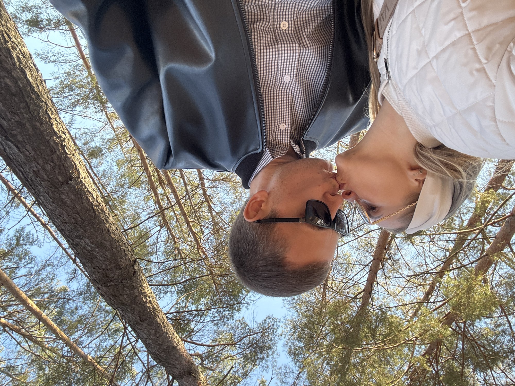 The image shows a couple kissing, shot from a low angle looking up. The man is wearing a black leather bomber jacket over a checkered shirt and sunglasses. The woman is wearing a white quilted jacket and a white headband. They are in a wooded area, with trees and branches visible against a blue sky. Given the request for a location in Chaikovsky, Russia, the scene likely depicts a park or forest area in the vicinity of the city. The act of kissing suggests an intimate relationship between the two people.