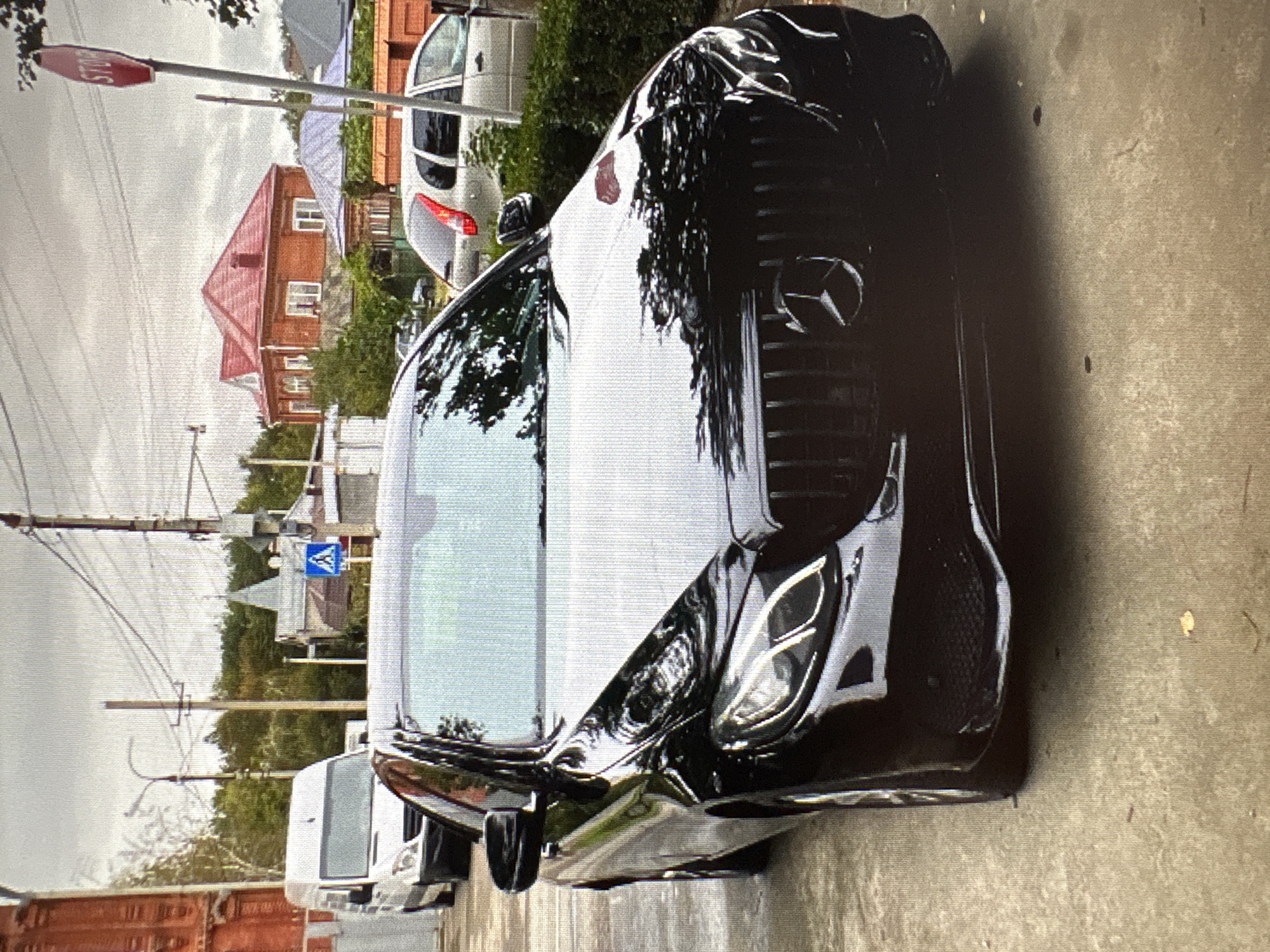 A black Mercedes-Benz sedan is parked on a paved surface, likely a street or driveway, with its front end angled towards the viewer. The car has a prominent grille with the Mercedes-Benz logo. In the background, a white cargo van is visible on the left, and a silver sedan is parked on the right. A residential building with a red tiled roof is prominent behind the silver car. A red circular "STOP" sign is mounted on a pole to the right of the frame. Overhead, power lines crisscross the cloudy sky. The overall environment suggests a moderately built-up urban or suburban area.